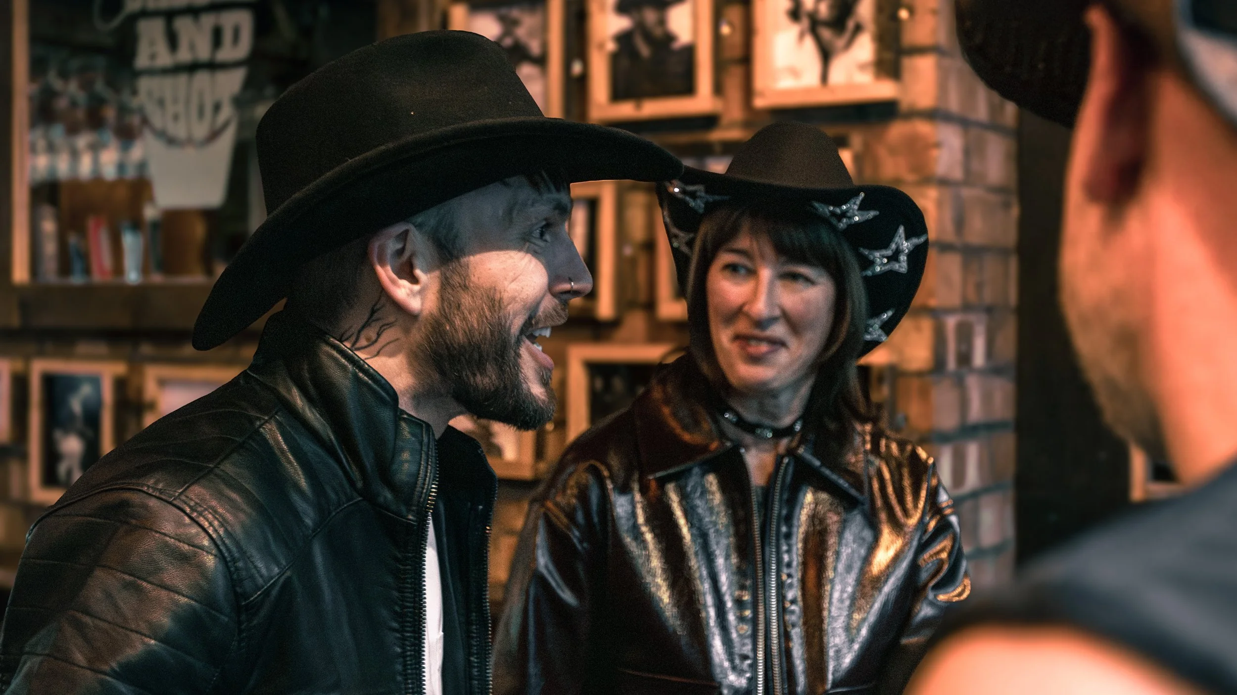 A man and woman dressed in dark clothing and black hats are talking and smiling in a cozy, dimly-lit bar or restaurant with brick walls and framed pictures in the background.