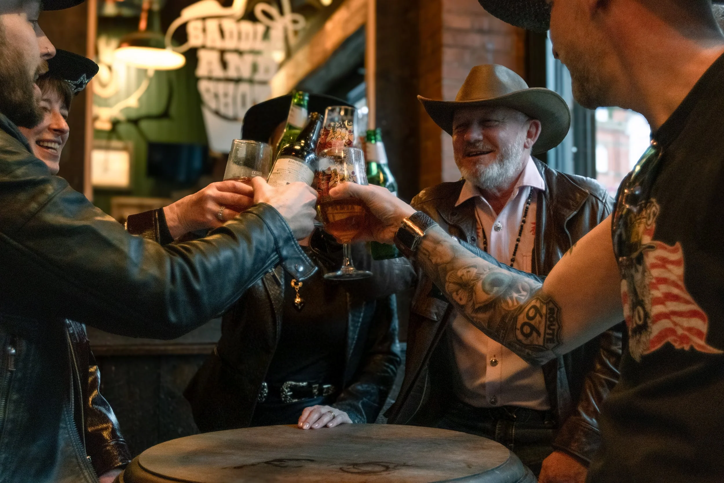 Group of people raising glasses of beer in a toast inside a bar, with some smiling and wearing casual clothing, including leather jackets and cowboy hats.