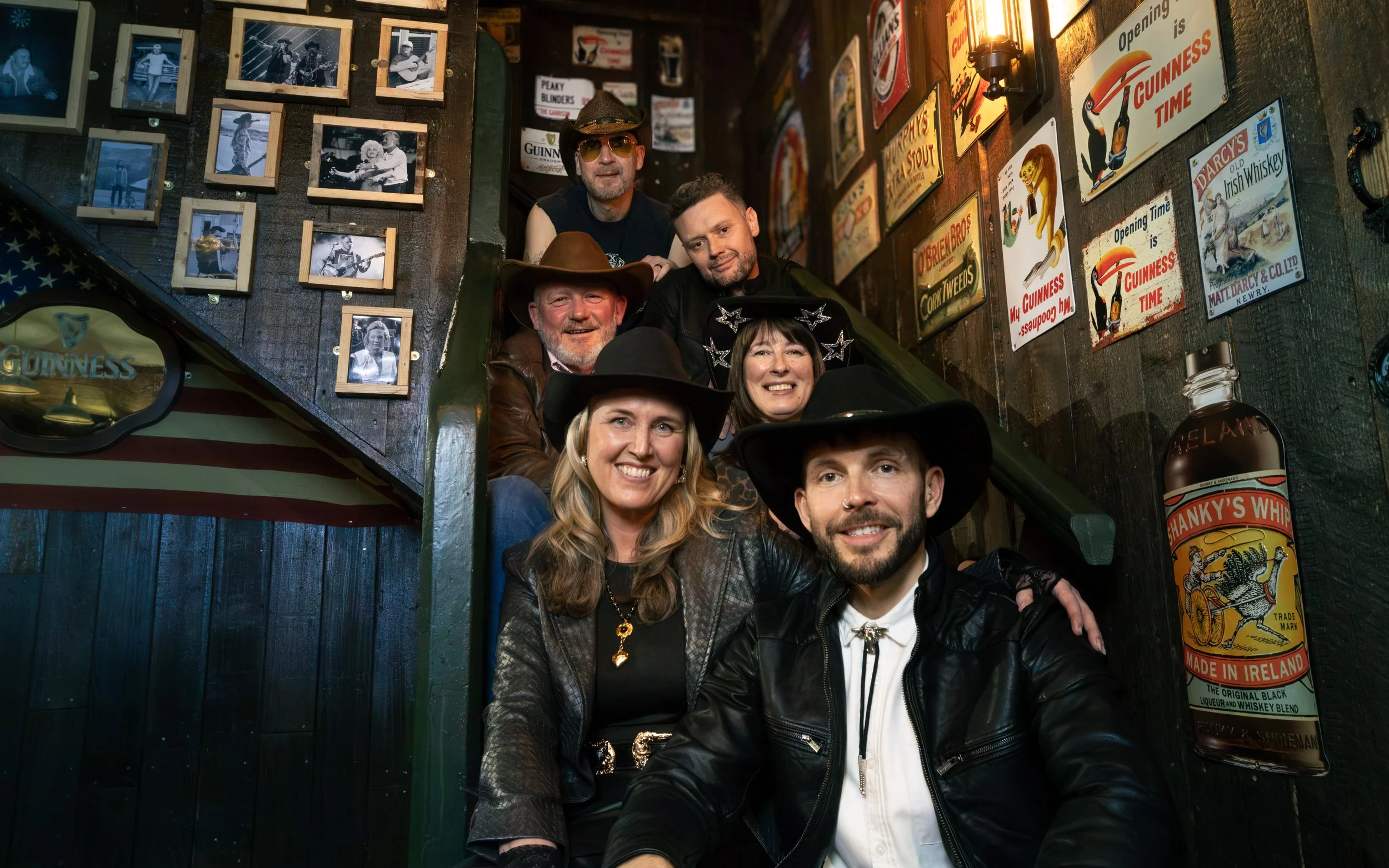 A group of six people, four women and two men, posing on a staircase inside a bar decorated with vintage signs and black-and-white photographs of musicians. The people are wearing casual clothing, some with cowboy hats and others with sunglasses. One