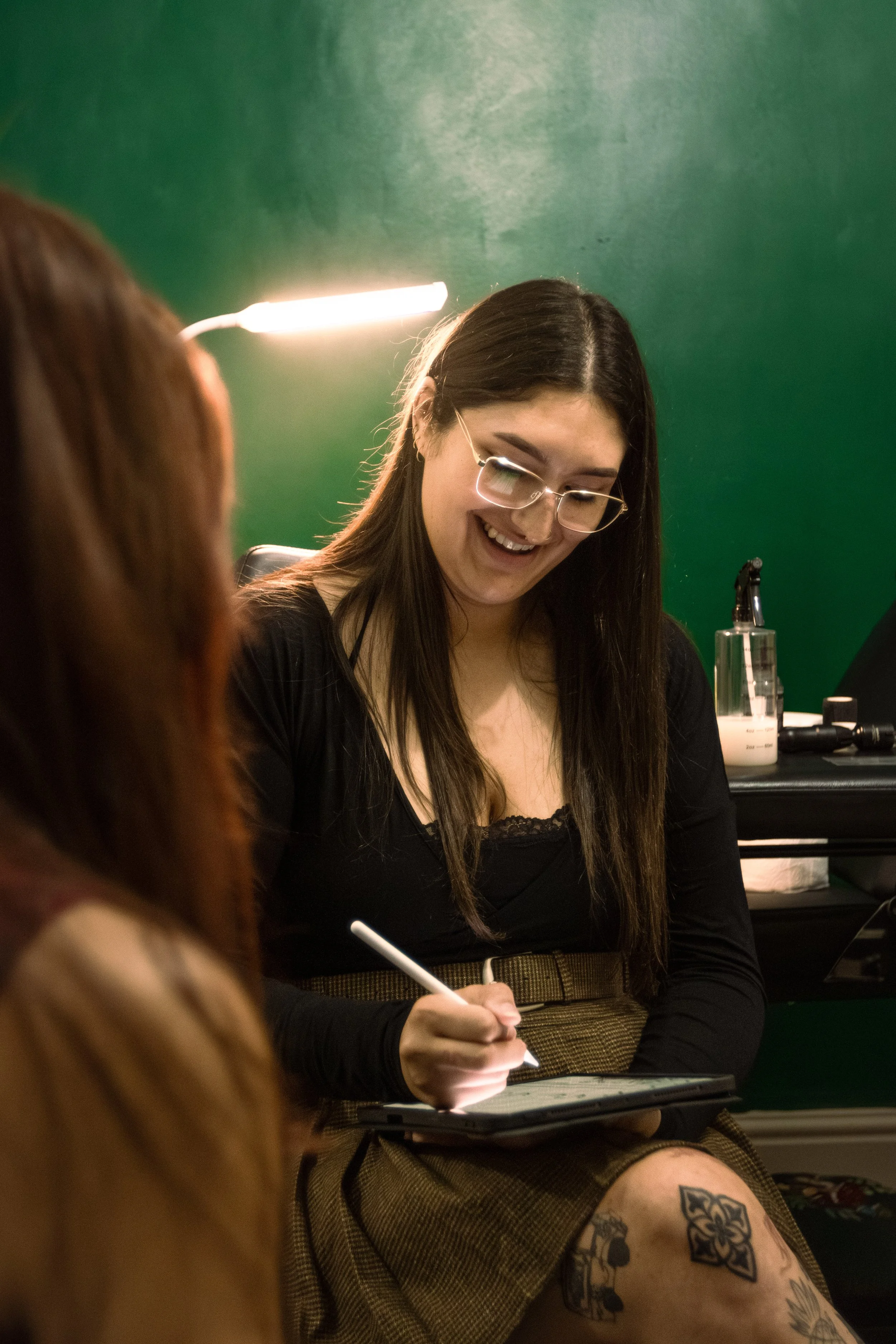 A woman with long dark hair, glasses, and tattoos on her legs, smiling while writing with a pen on a notepad during a tattoo session in a studio with green walls.