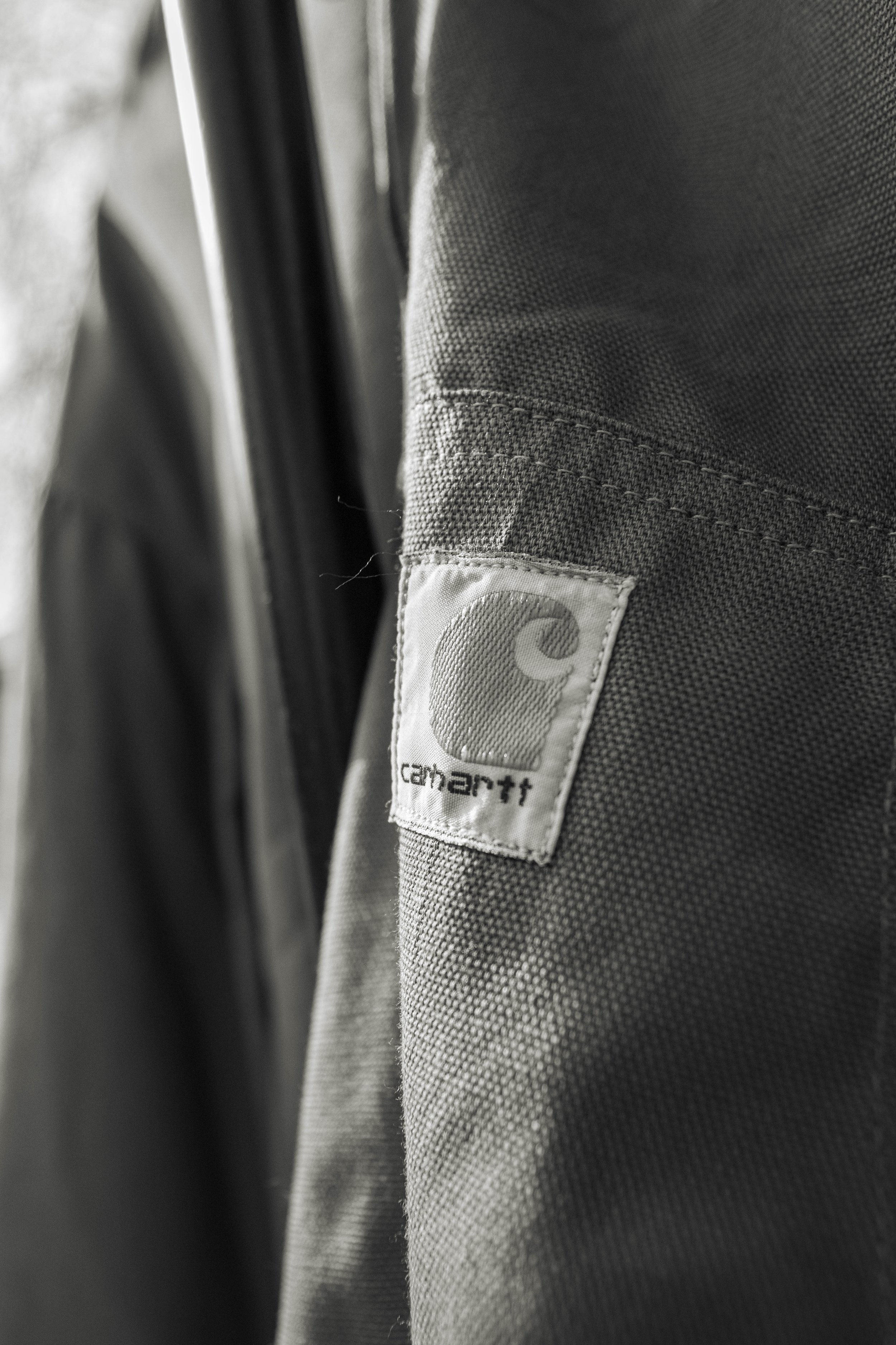 Close-up of a gray Carhartt work jacket hanging on a black hanger, with a focus on the Carhartt label on the fabric.