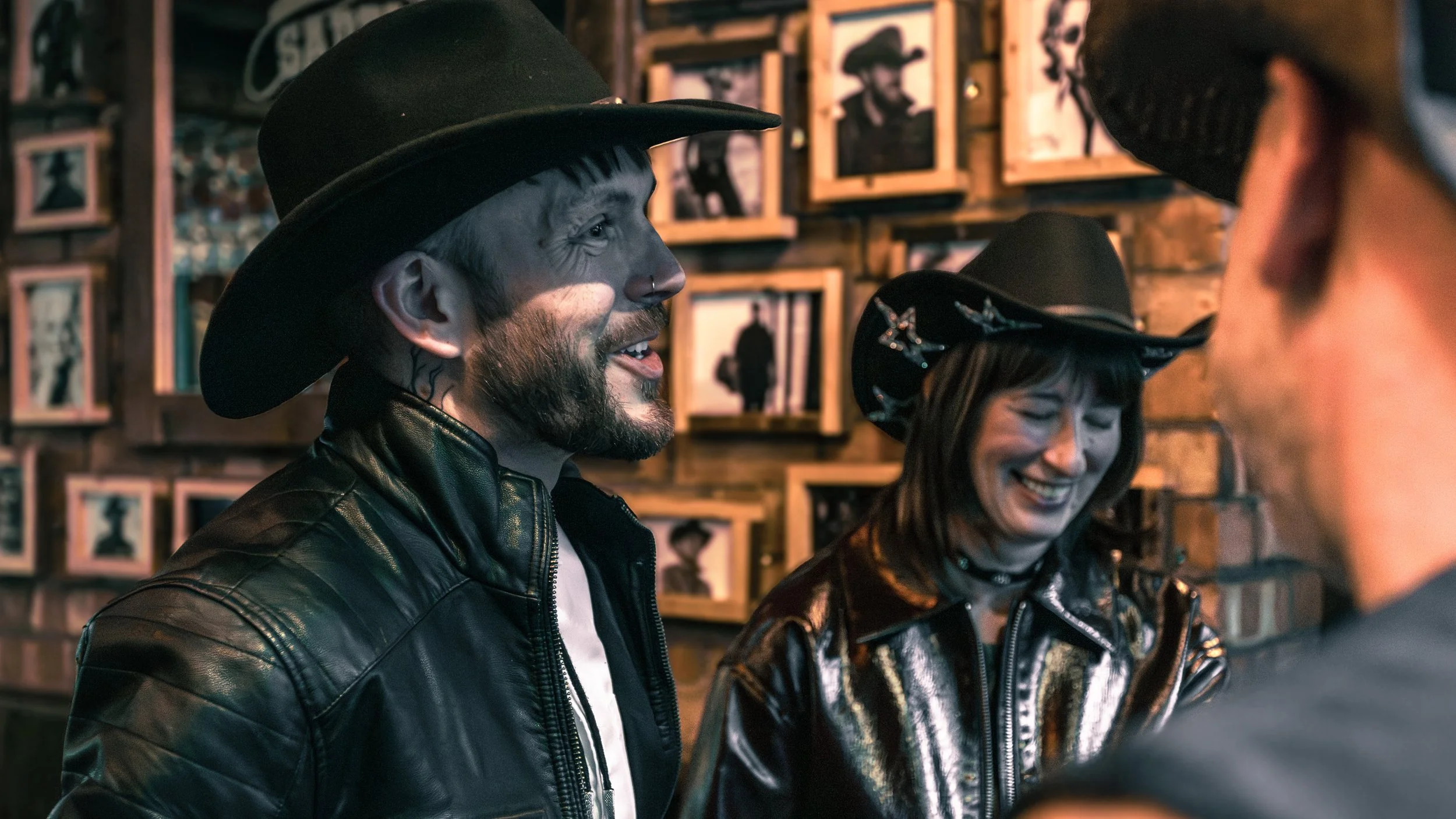 Two people, a man with face paint and a woman, wearing cowboy hats and leather jackets, laughing and talking in a bar with framed photos on the wooden wall behind them.