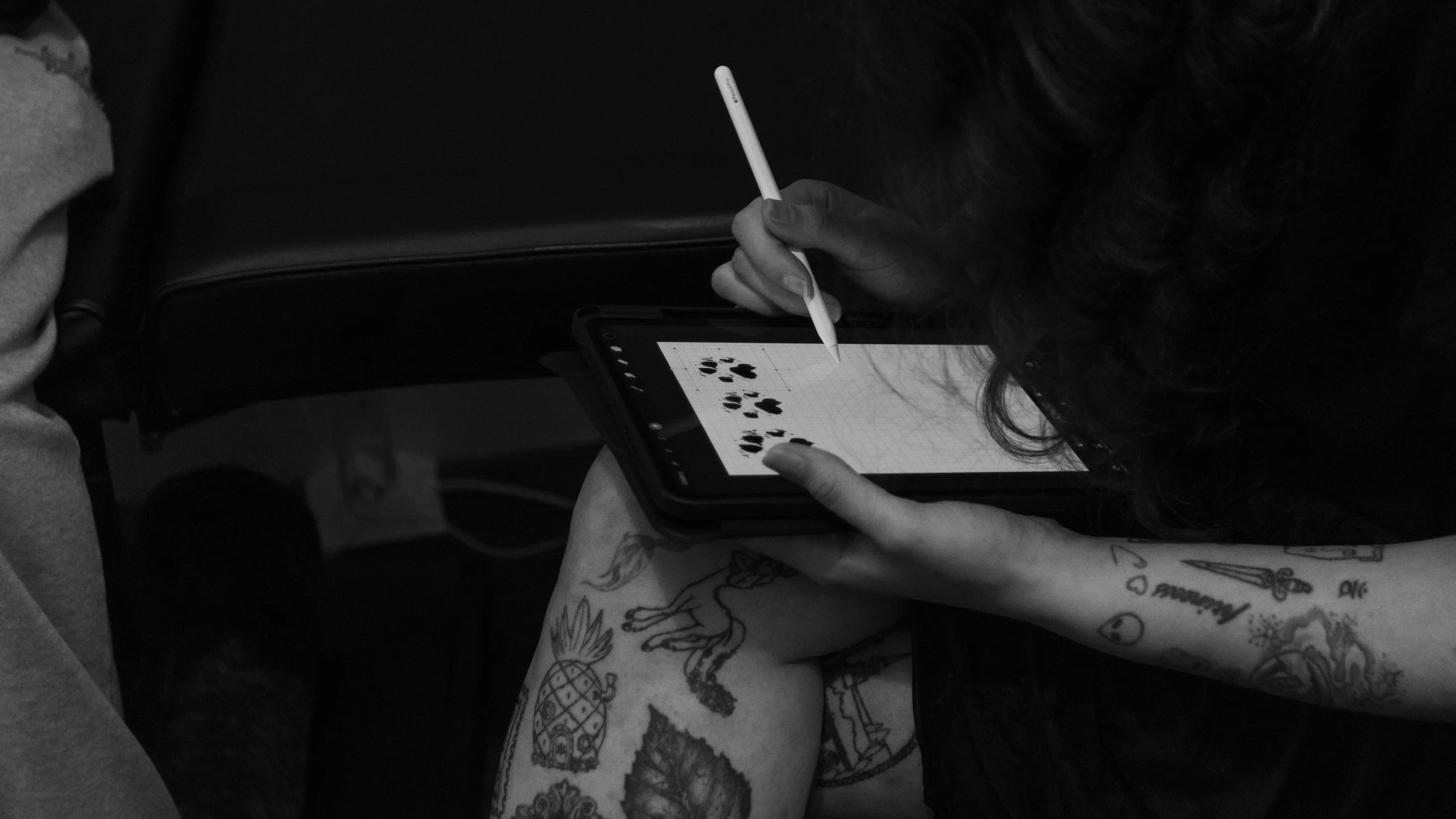 A person with tattoos on their arm using a stylus to draw black ink paw print patterns on a tablet.