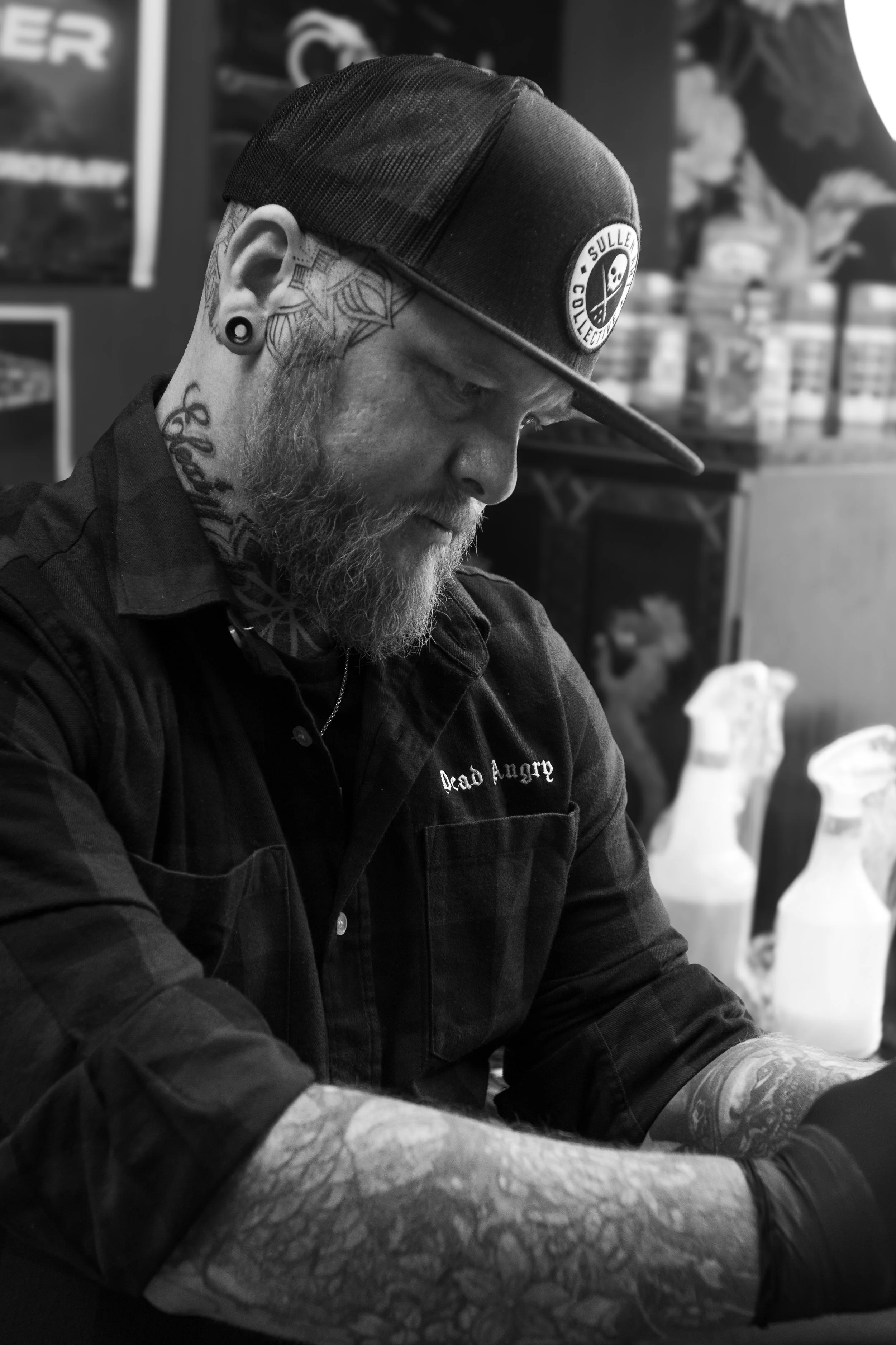 A man with tattoos and gauges in his ears, wearing a black mesh trucker hat with a circular patch, is sitting at a table. He has a beard and is wearing a black shirt. The background includes shelves with products and a blurred figure, suggesting a st