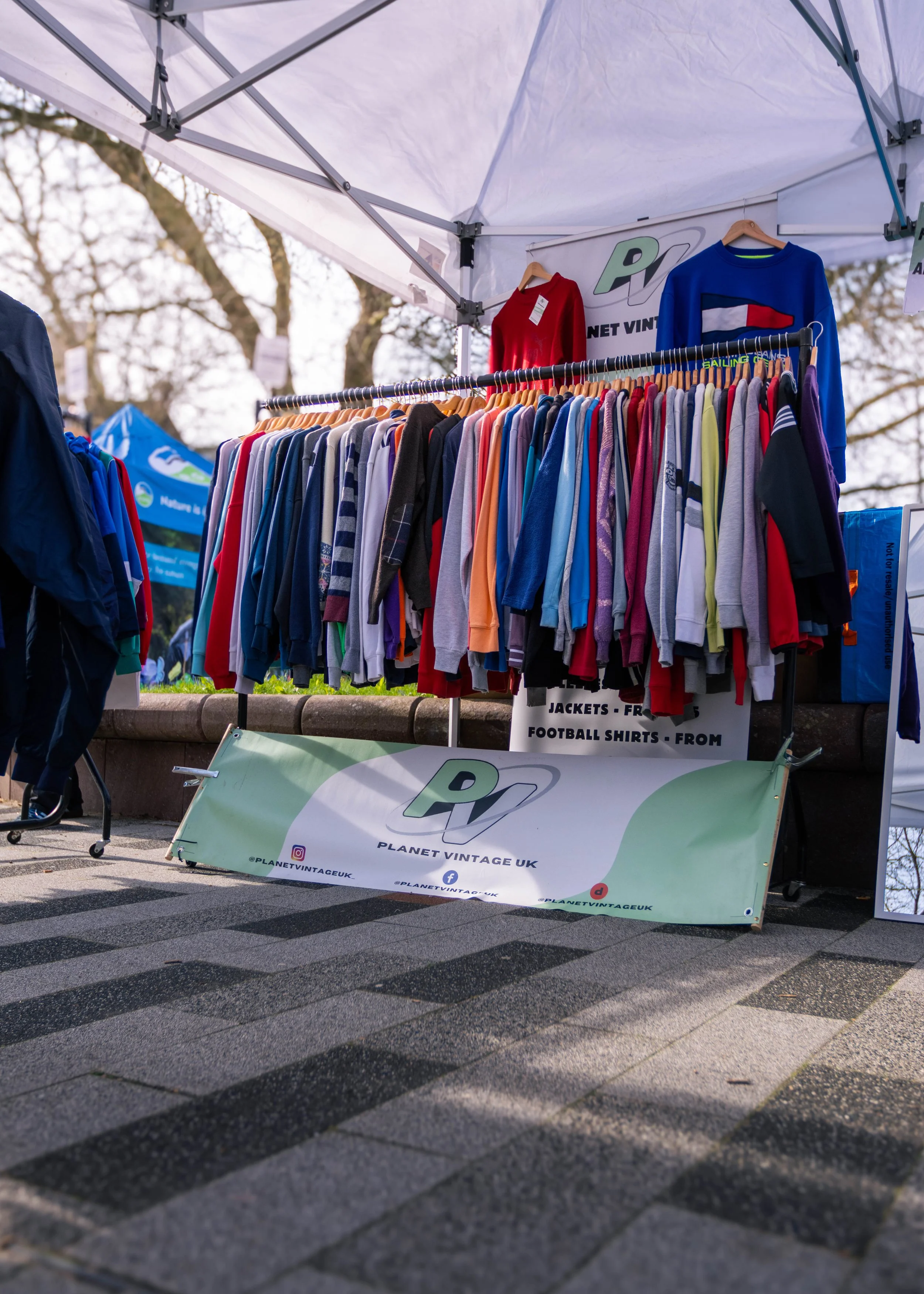 A clothing stall with a variety of sports jackets and shirts hanging on a rack under a white canopy. The stall has a banner with 'Planet Vintage UK' and social media icons.