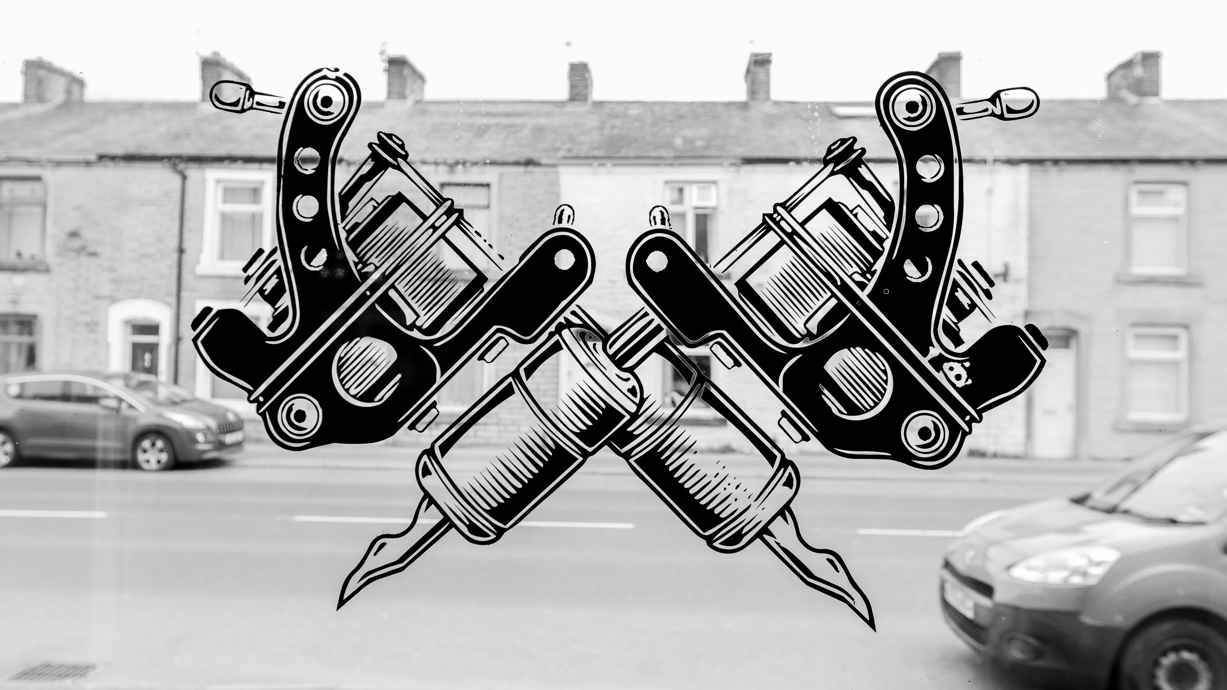 Black and white graphic of crossed handguns with a skull in a heart at the bottom, superimposed on a window reflecting a street scene with cars and houses.