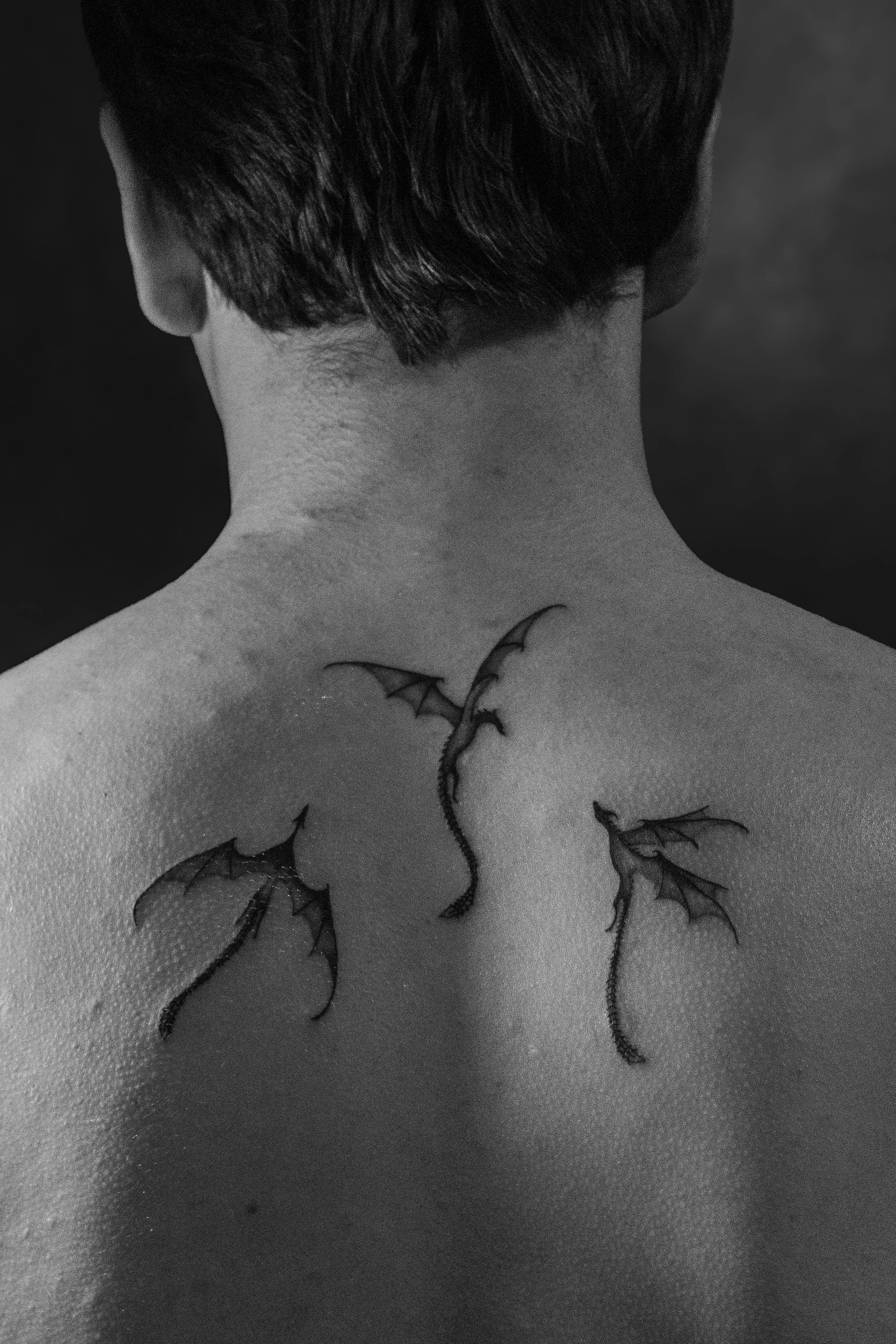 Back view of a person with short dark hair, featuring a dragon tattoo on the upper back and neck, with a dark, blurry background.