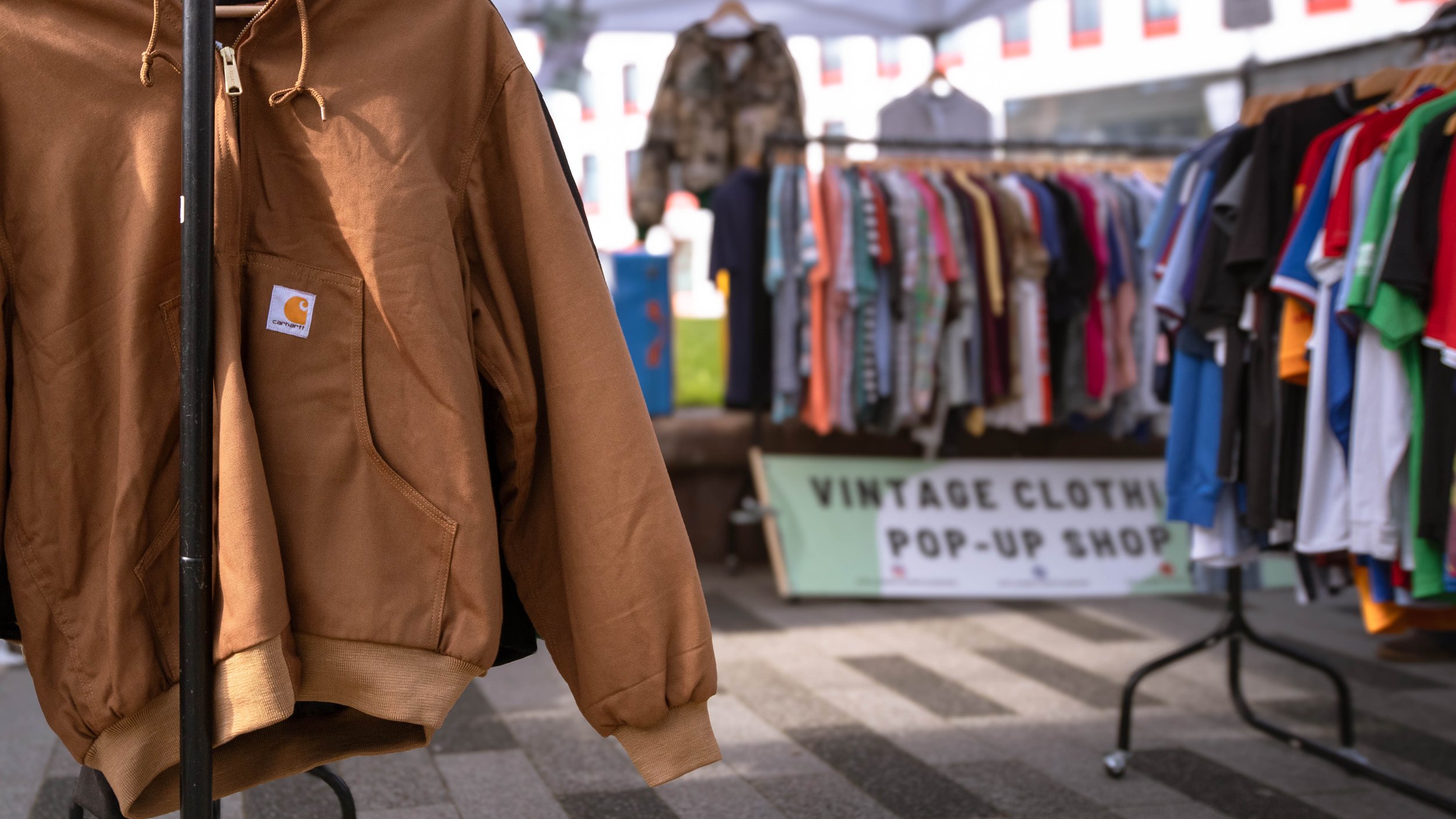 Close-up of a person wearing a brown Carhartt jacket, standing at a vintage clothing pop-up shop. In the background, a rack of colorful clothes and a sign reading 'VINTAGE CLOTHING POP-UP SHOP' are visible.
