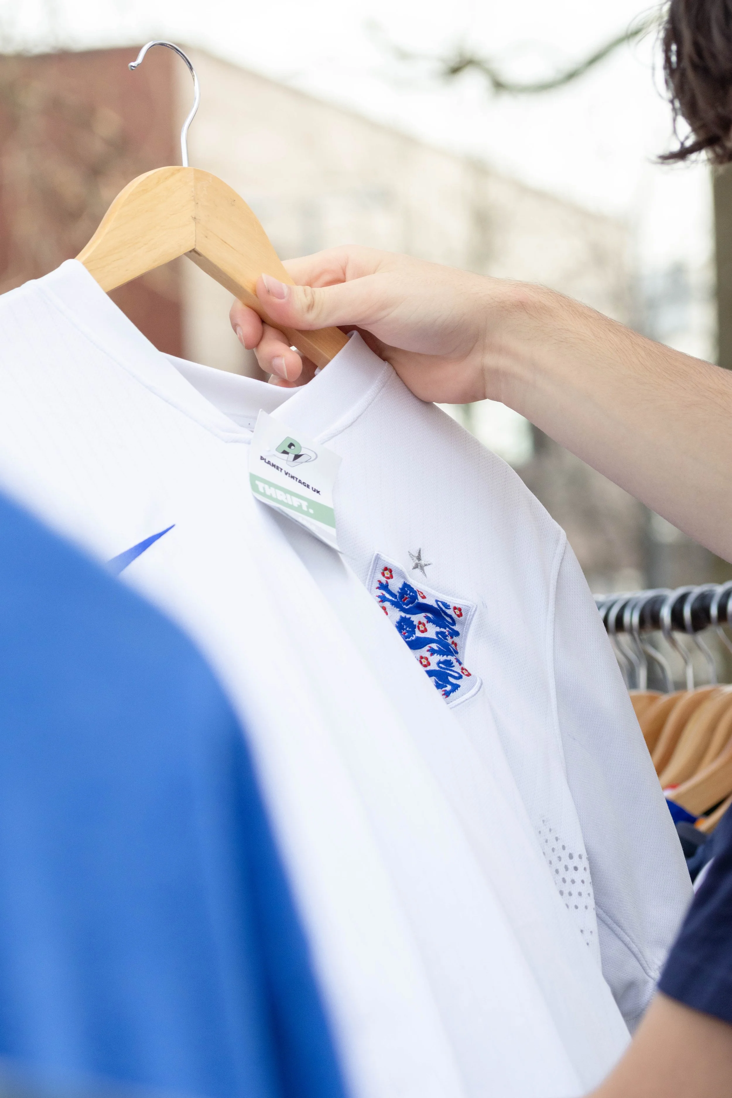 A person holding a white football jersey with England cricket team's emblem on it, hanging on a wooden hanger in an outdoor setting.