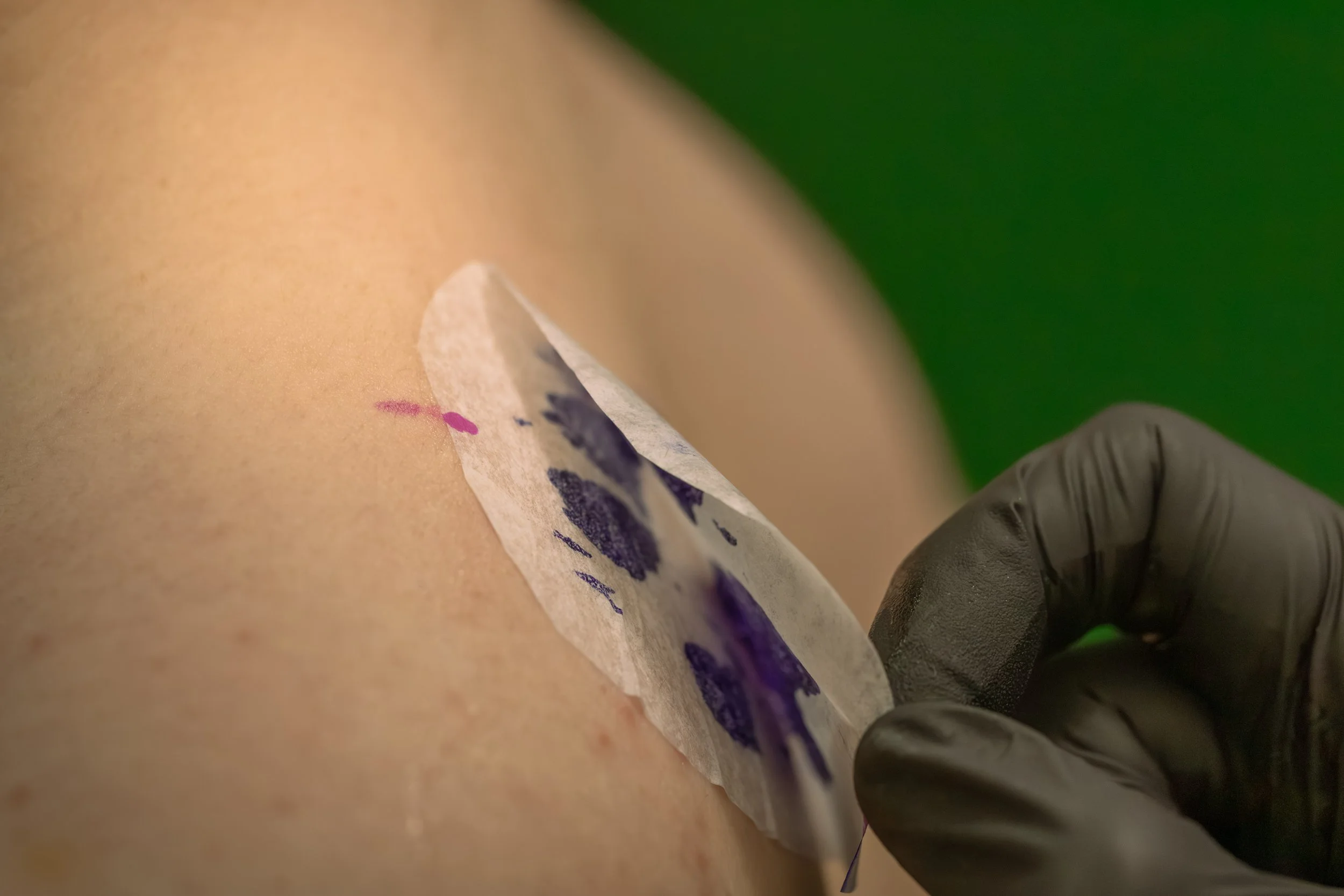 Close-up photo of a tattoo being inked on skin, with a tattoo needle and ink visible, and a person wearing black gloves holding the needle.