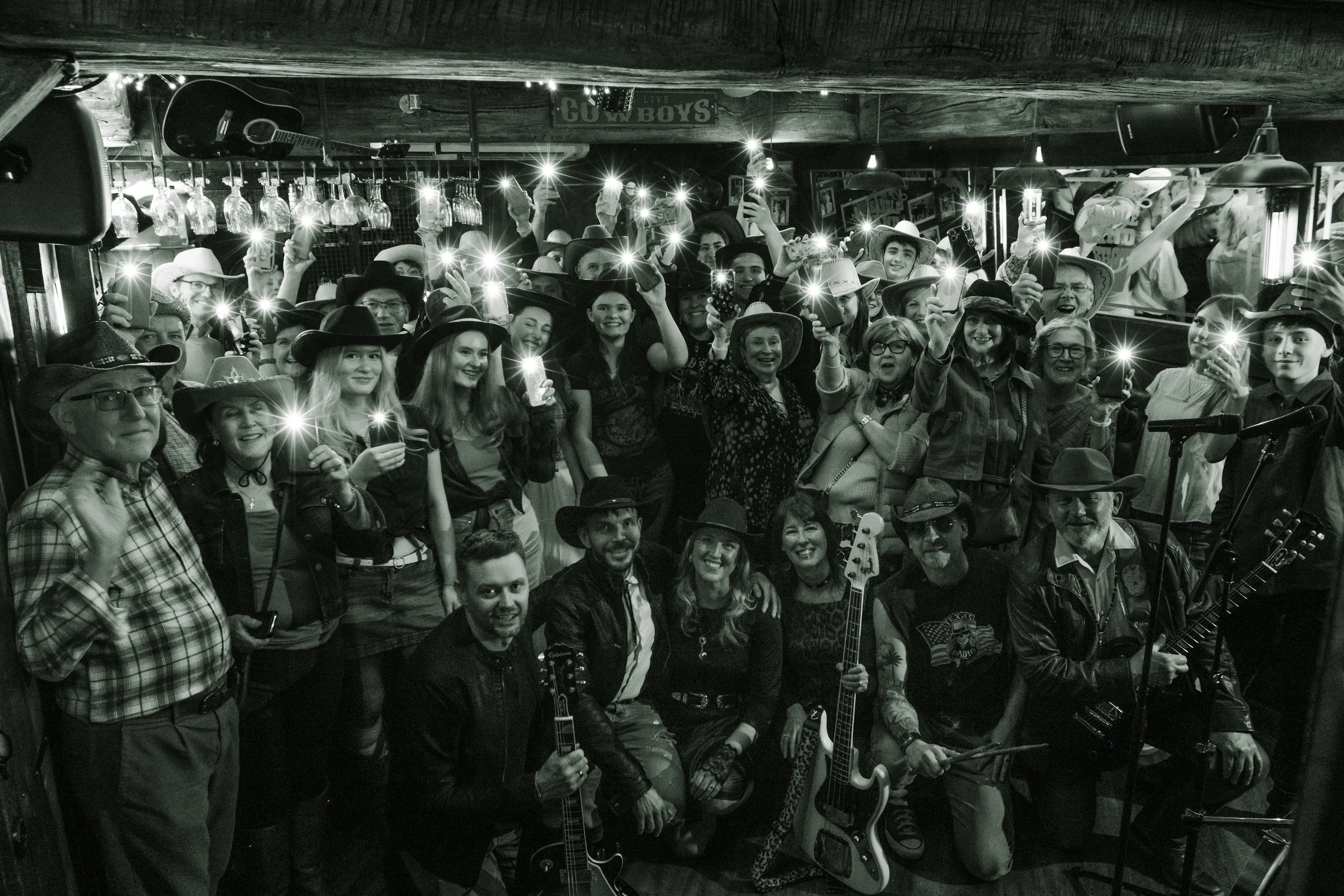 A large group of people, many wearing cowboy hats, gather in a bar or restaurant with musical instruments, including guitars. Some are holding drinks and phones with flashlights, smiling and posing for the photo. The venue has wooden decor and hangin