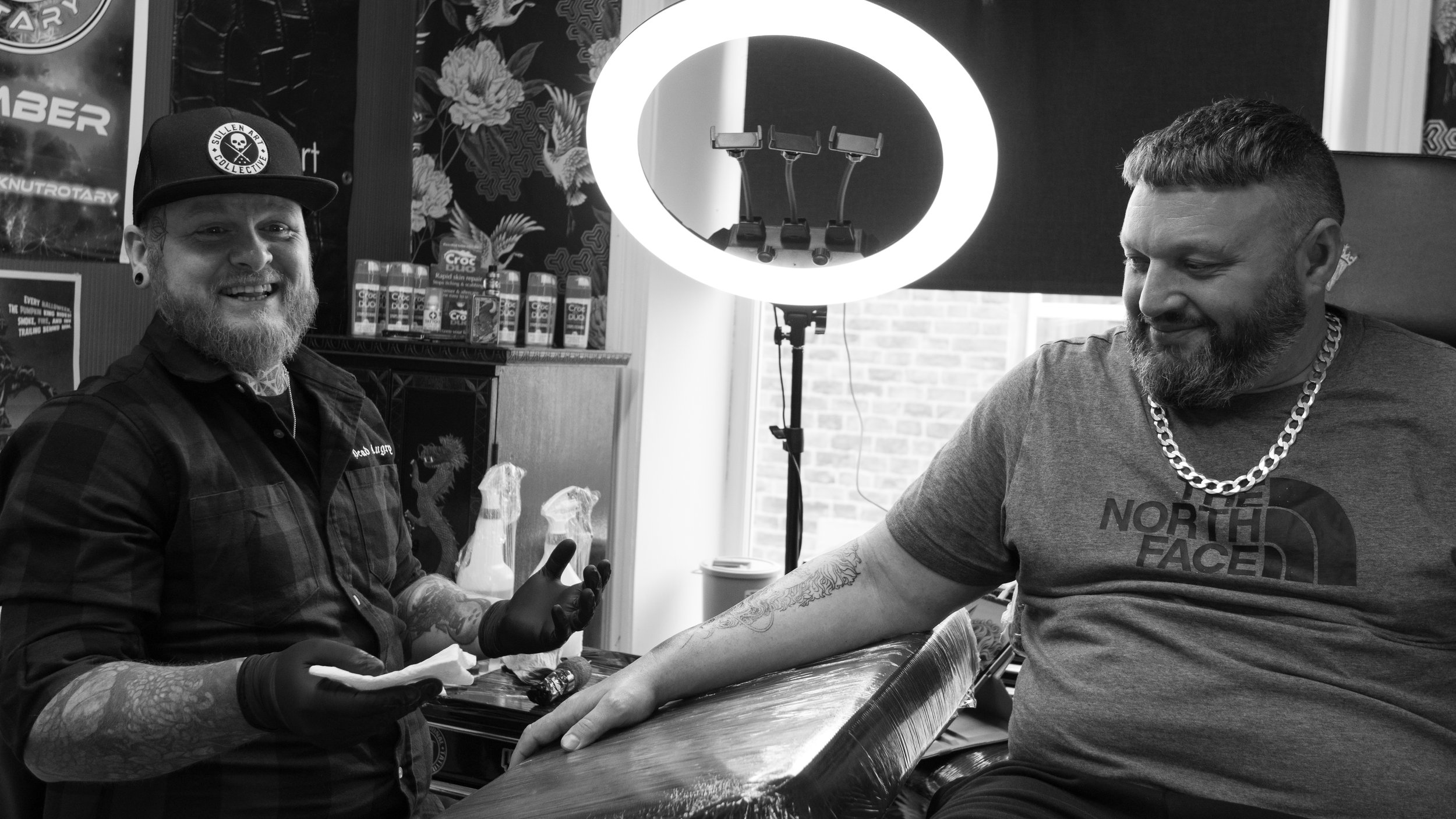 Two men with tattoos and beards, one wearing a North Face shirt and the other wearing a cap with a skull logo, are in a tattoo shop. The man on the right is getting a tattoo on his arm while the man on the left is smiling and wearing black gloves. A 