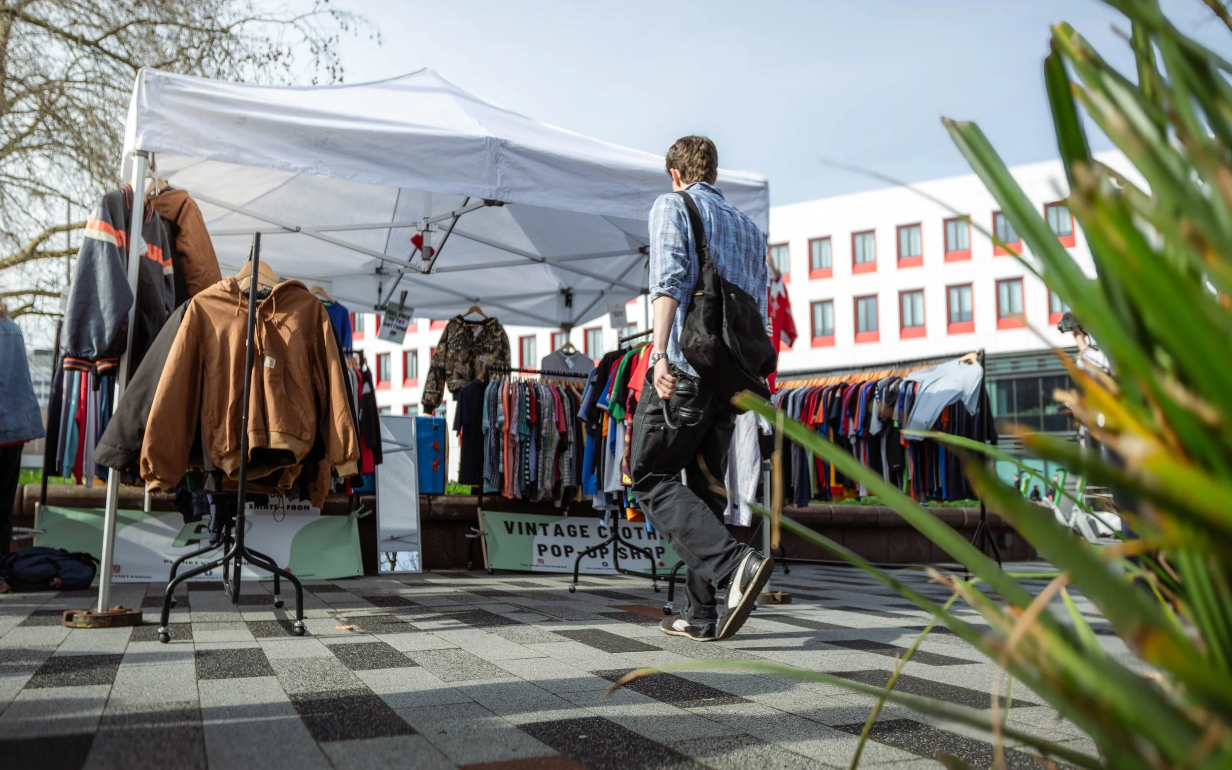 A person shopping at an outdoor vintage clothing pop-up shop, browsing racks of clothes under a white canopy, with a modern building in the background.