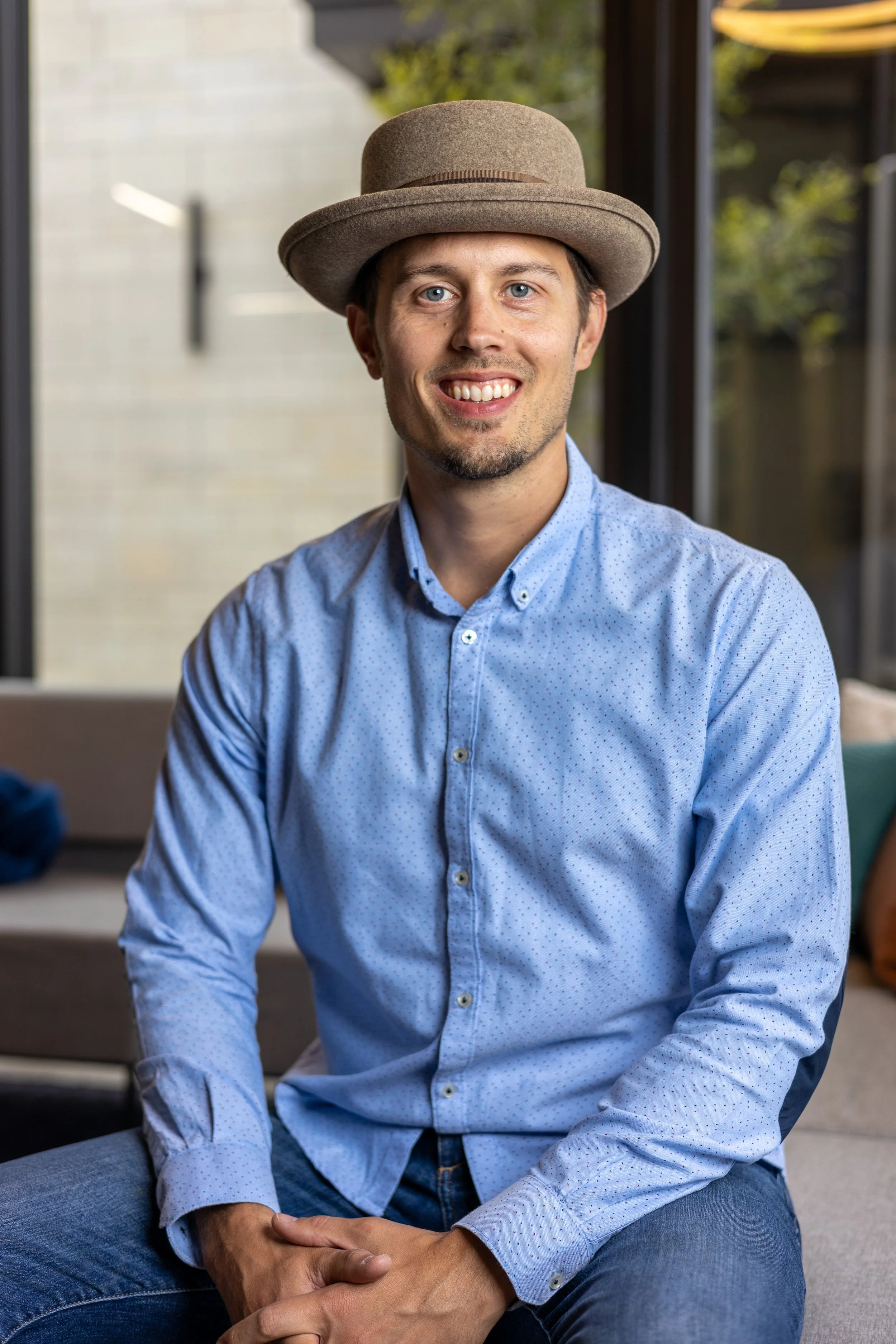 A smiling man wearing a light blue button-up shirt and a beige fedora hat, sitting indoors with a blurred background of windows and greenery.