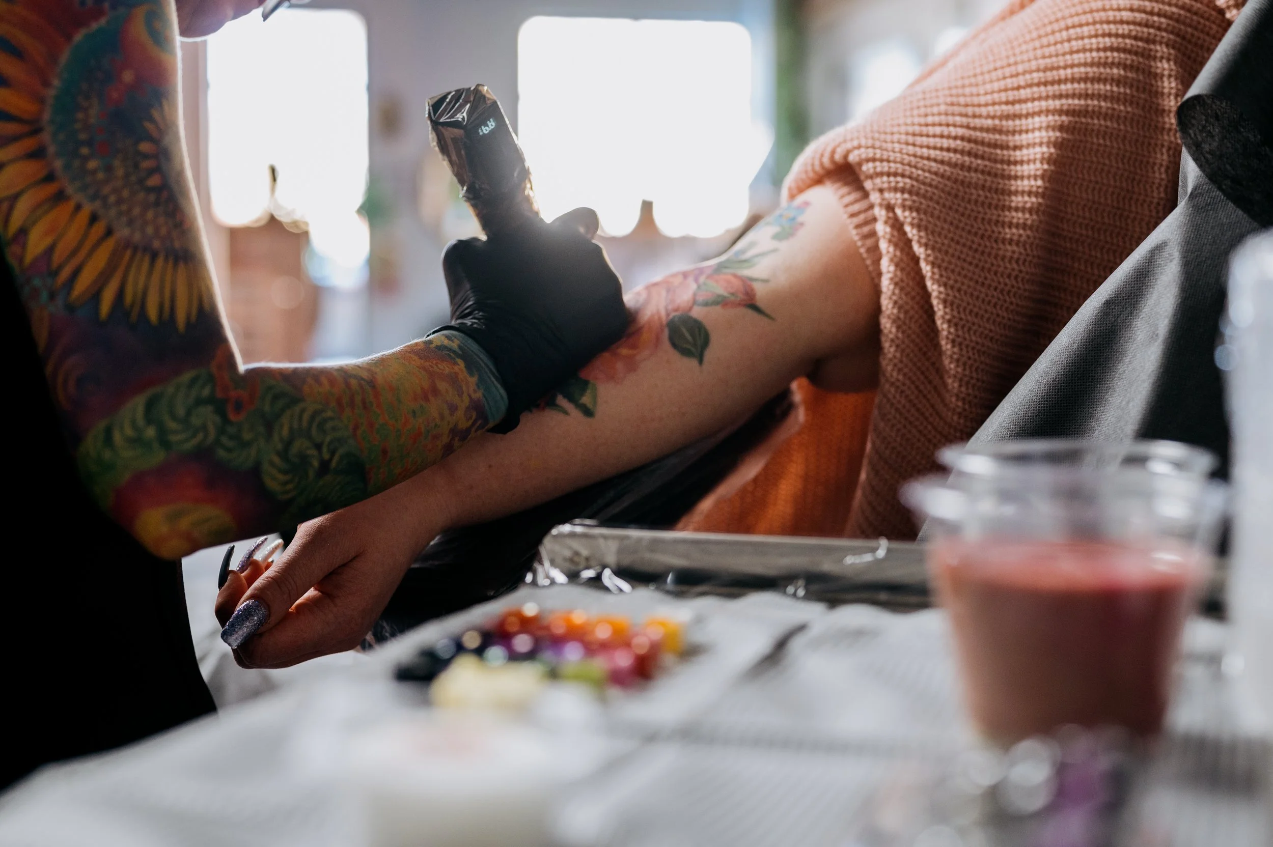 A tattooed person with black gloves holding a tattoo machine and tattooing the arm of another person wearing a coral-colored sweater, with tattoo supplies on a table in the foreground.