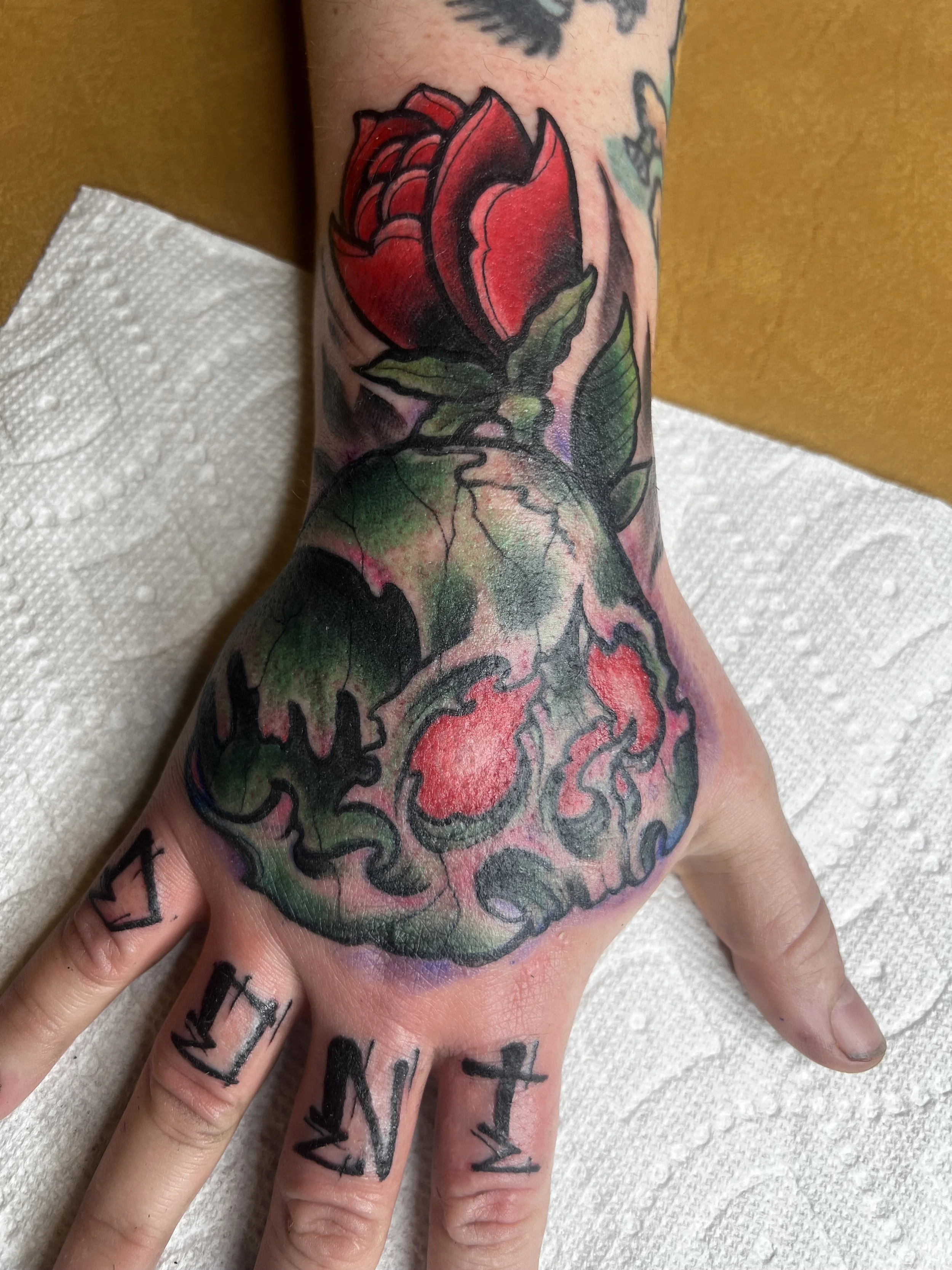 A colorful tattoo of a red rose with green leaves on a person's hand and wrist.