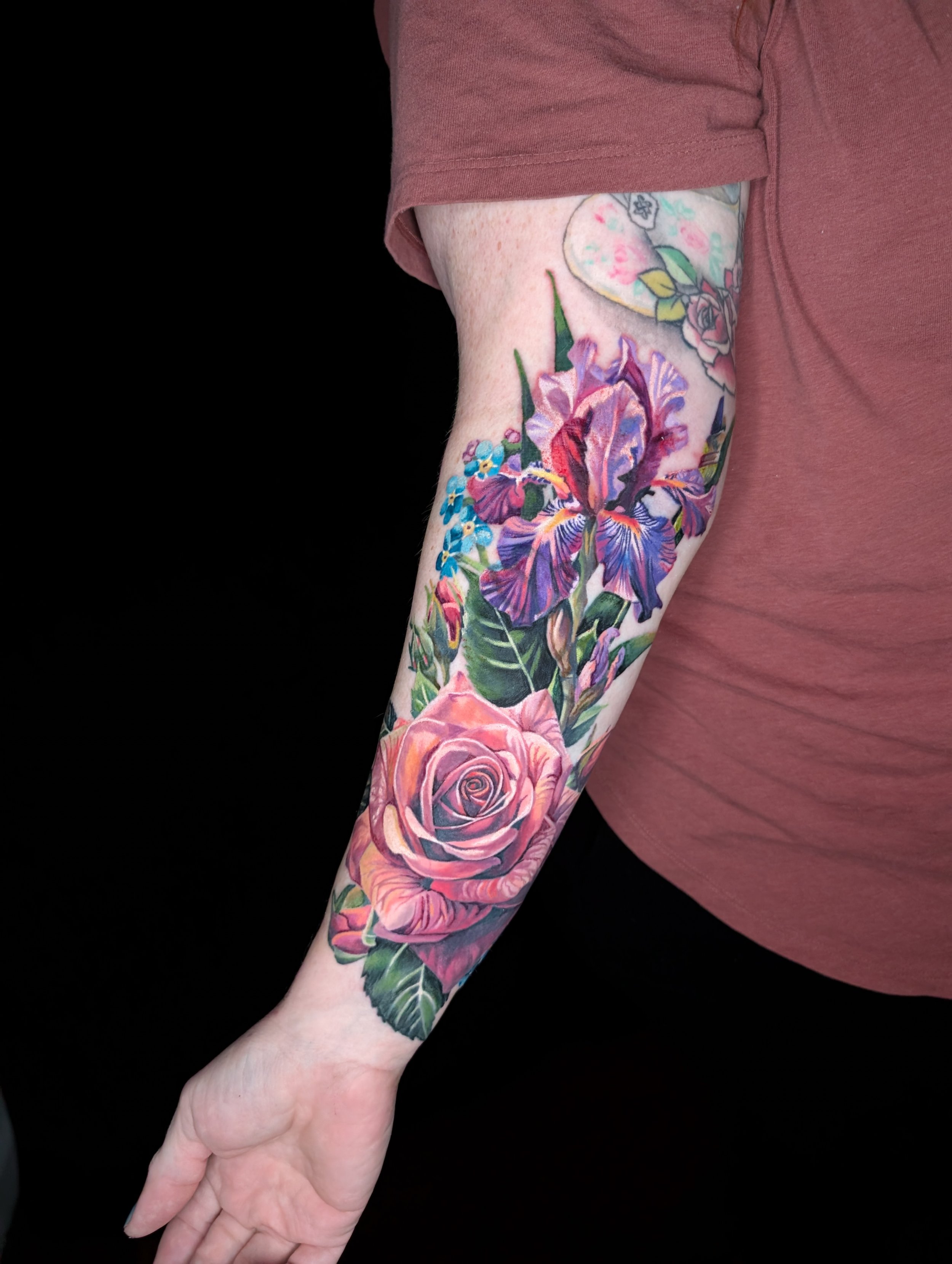 A person's arm and hand with a floral tattoo featuring large pink, purple, and blue flowers, against a black background with a brown shirt sleeve.