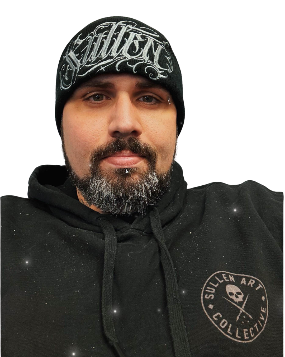 Close-up of a man wearing a black beanie with intricate white embroidery, a black hoodie with a "Sullen Art Collective" logo featuring a skull and a paintbrush, and with facial hair and an eyebrow piercing.