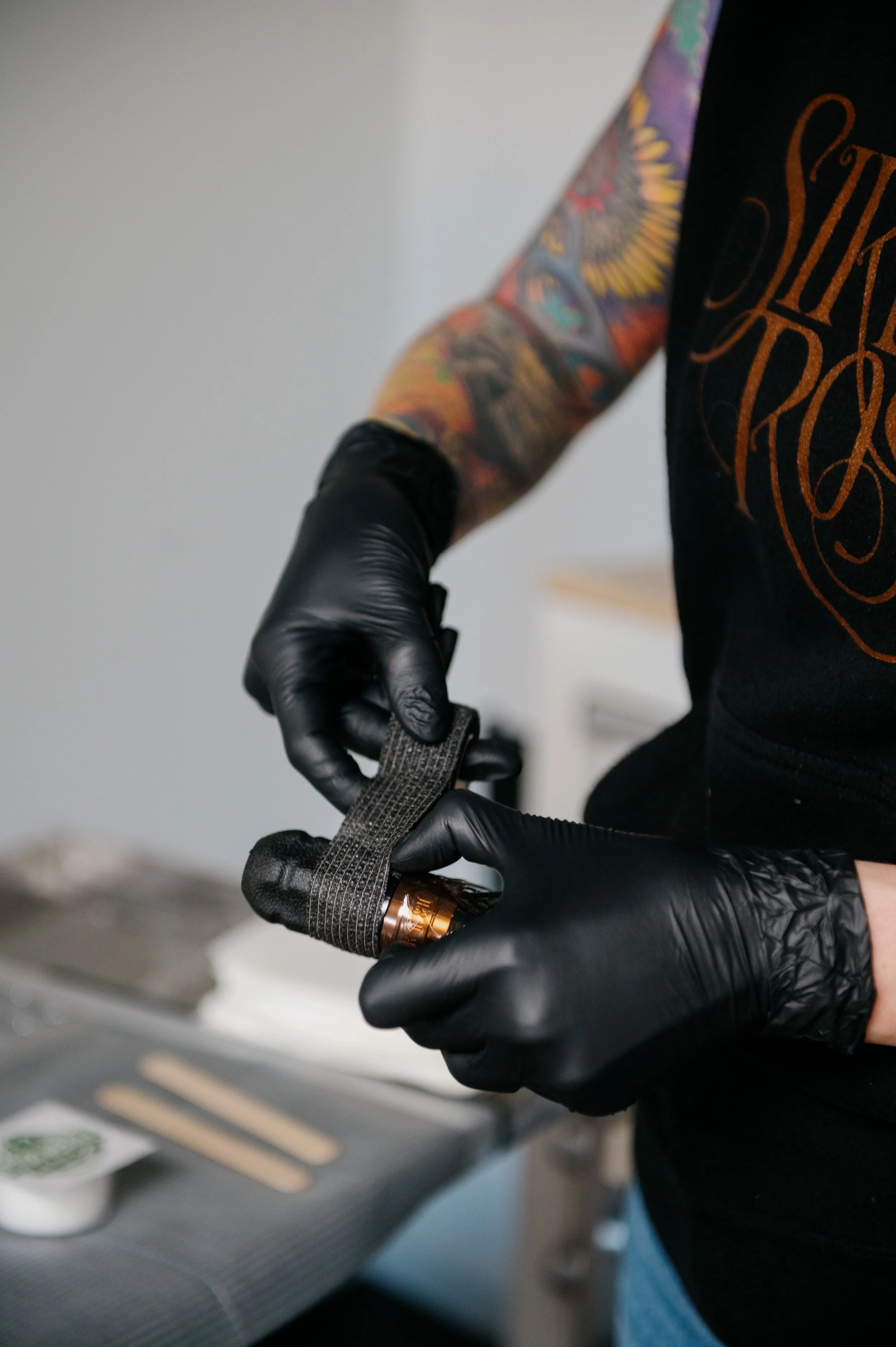 A person with a colorful tattoo sleeve on their arm wearing black gloves. They are holding a tattoo machine, and part of their black shirt with orange lettering is visible. The background appears to be a tattoo studio or workspace.
