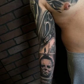Close-up of a person's arm with multiple tattoos, including a portrait of a man with glasses, a cityscape, and other designs, against a brick wall background.