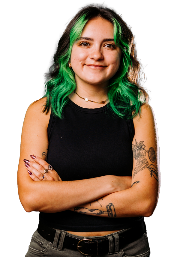 A young woman with green hair, wearing a black sleeveless top, standing with her arms crossed, smiling, with visible tattoos on her right arm, and jewelry including rings, a necklace, and earrings.