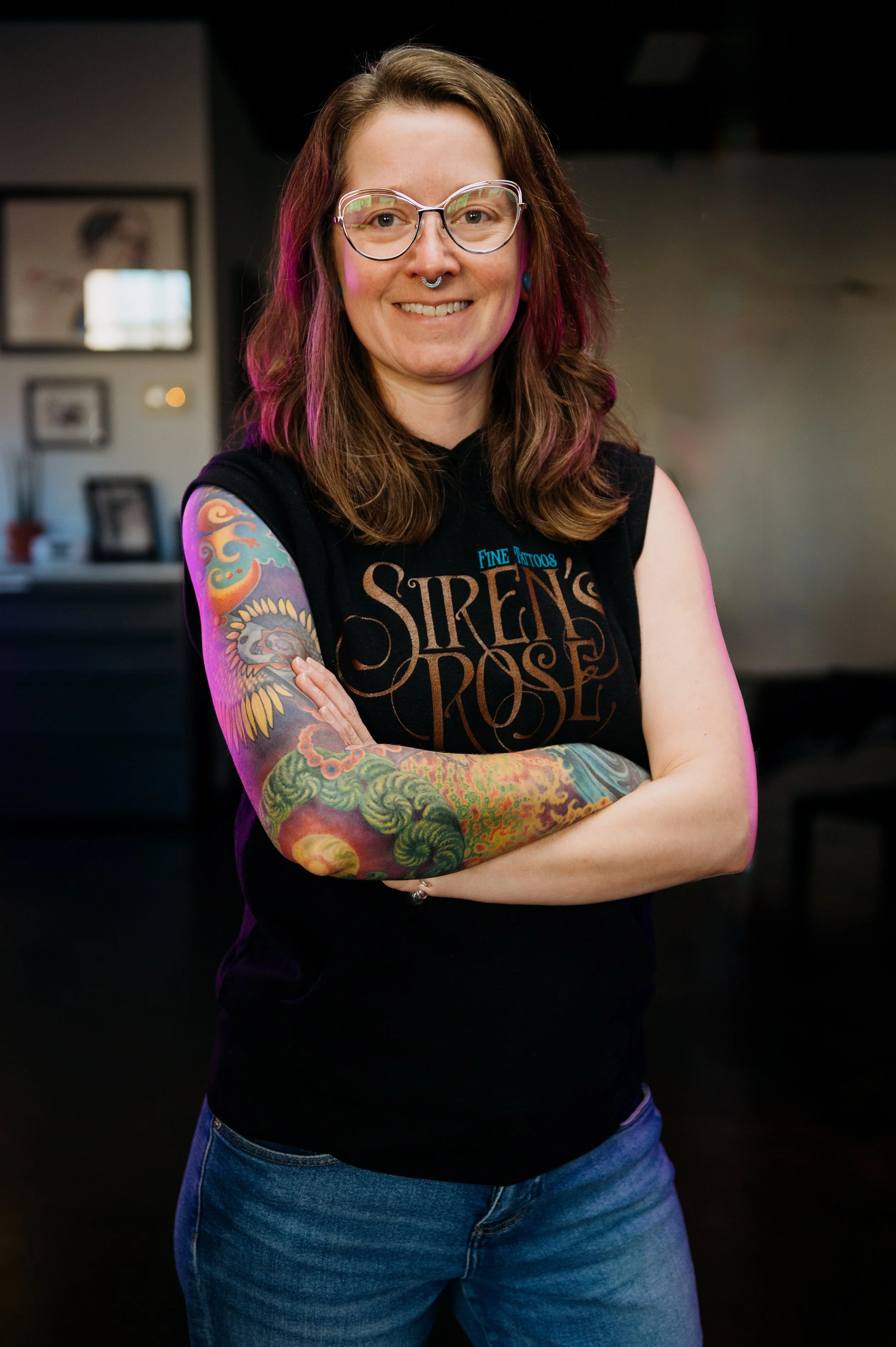 A woman with shoulder-length brown hair, wearing glasses, a septum piercing, and colorful tattoos on her arms, smiling and crossing her arms. She is wearing a sleeveless black shirt with text and blue jeans in a dark indoor setting.