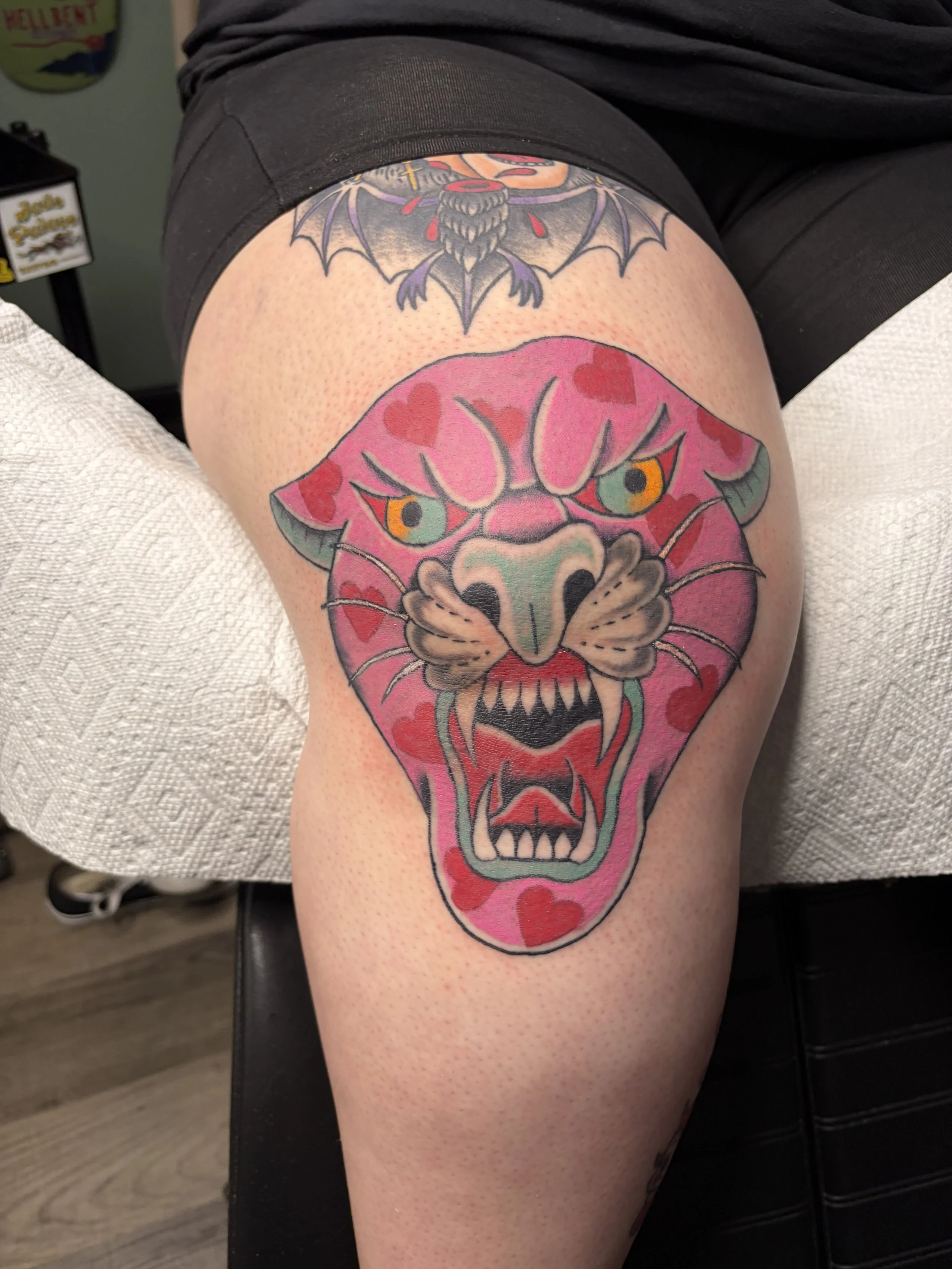 Tattoo of a fierce pink face with green eyes, adorned with red hearts and a dragonfly on its nose, on a person's upper thigh.