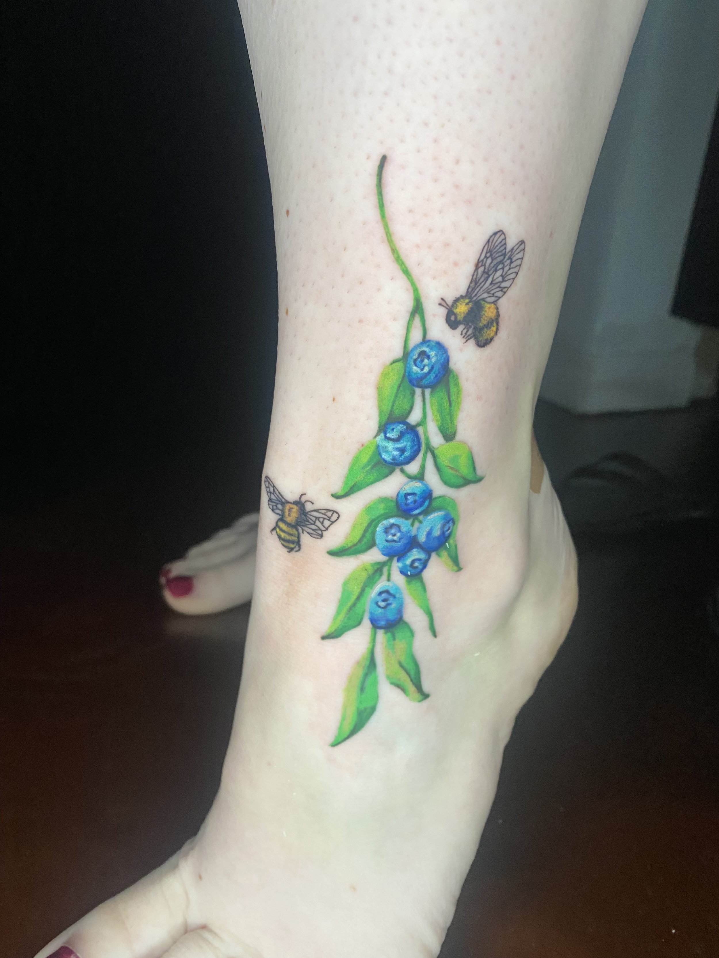 A colorful tattoo of a blueberry branch with leaves and two bees on a person's forearm.