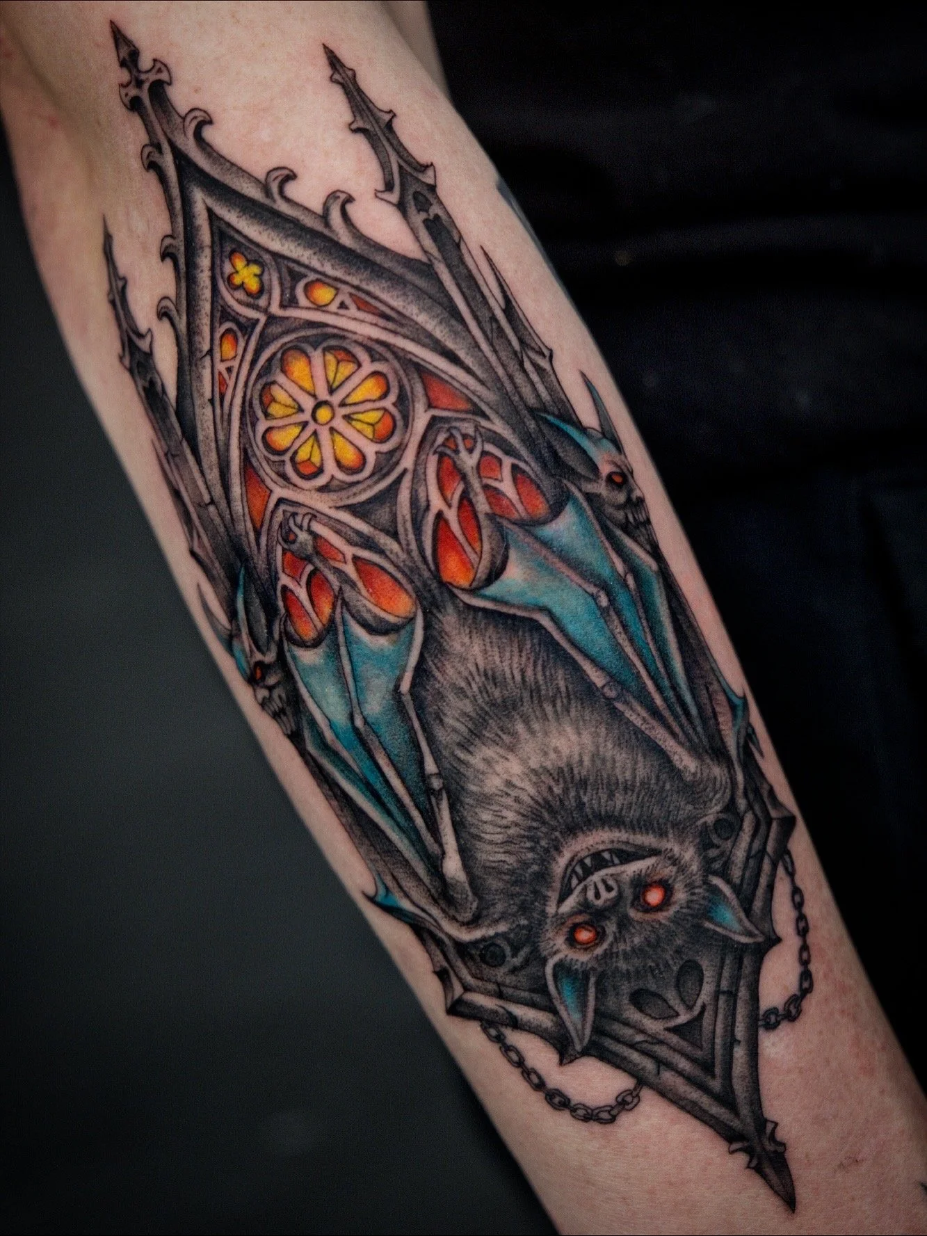 Tattoo of a bat with glowing eyes, surrounded by Gothic-style architectural designs and a skull, on a person's arm.