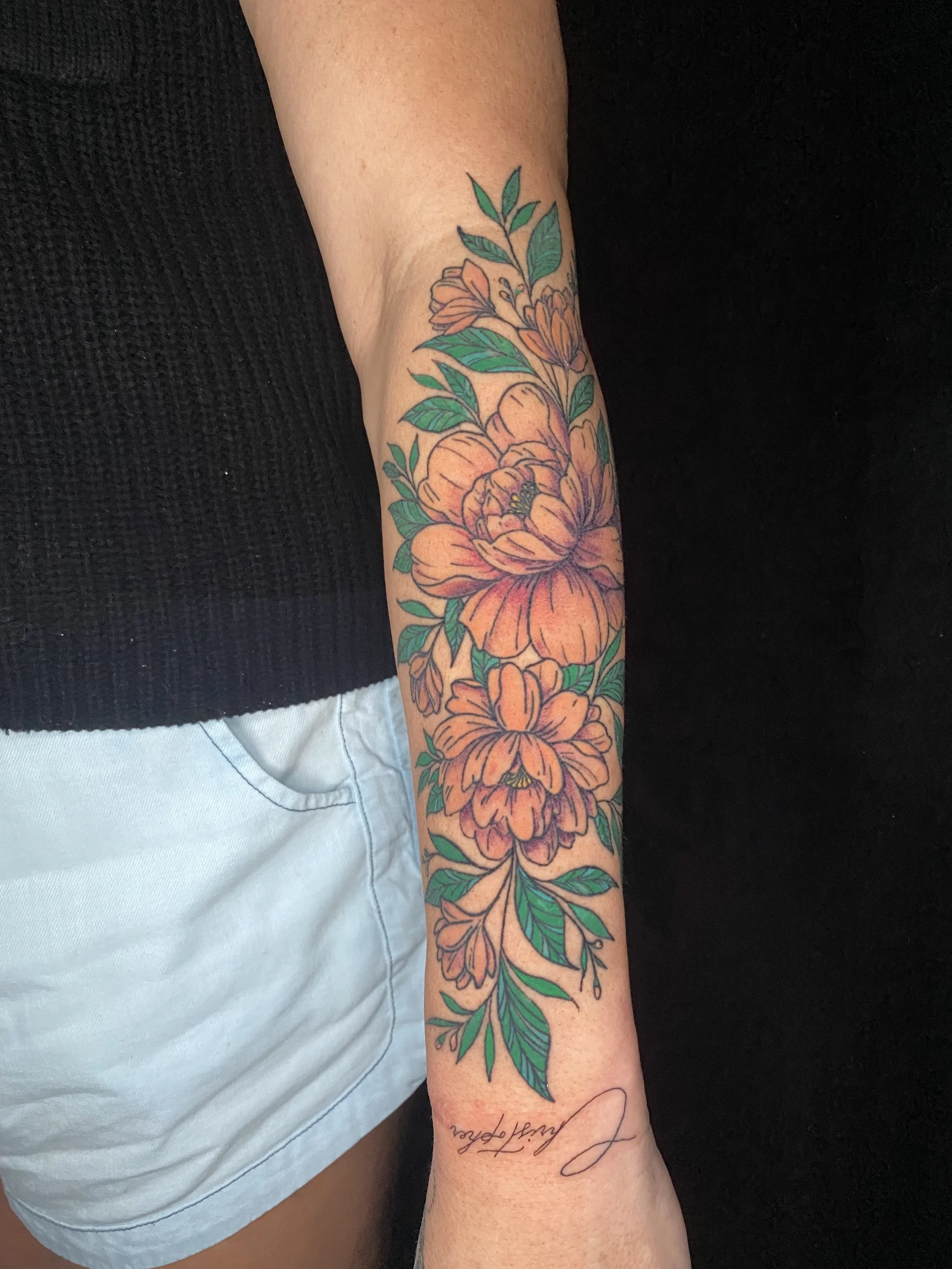 A person with a floral tattoo on their forearm featuring orange flowers and green leaves, with a cursive script tattoo below that reads 'Laughter'.