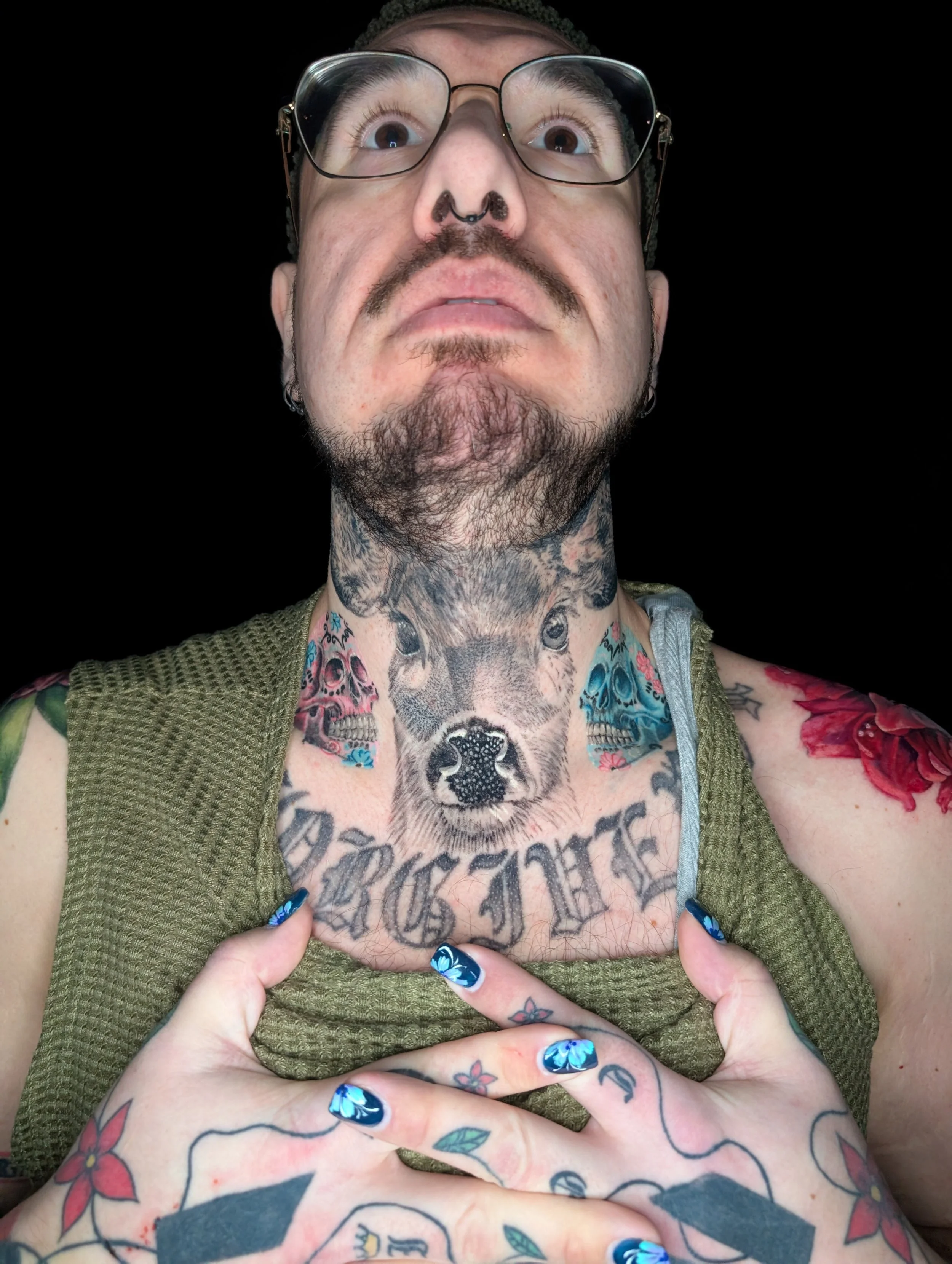 A man with glasses and tattoos exposing his chest, which has a large tattoo of a dog's face and surrounding gothic script. His hands, with painted nails, are holding his chest open. He has facial hair and piercings, and is looking up against a black background.