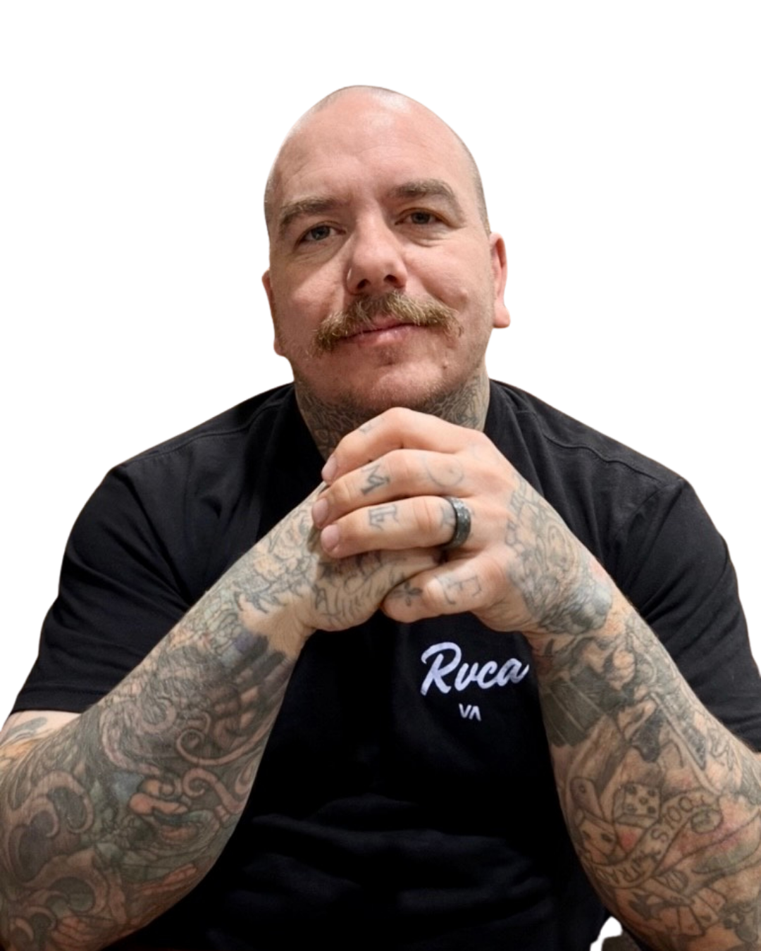 A man with a mustache and tattoos on his arms and hands, wearing a black t-shirt with white text, sitting with his hands clasped and looking at the camera.