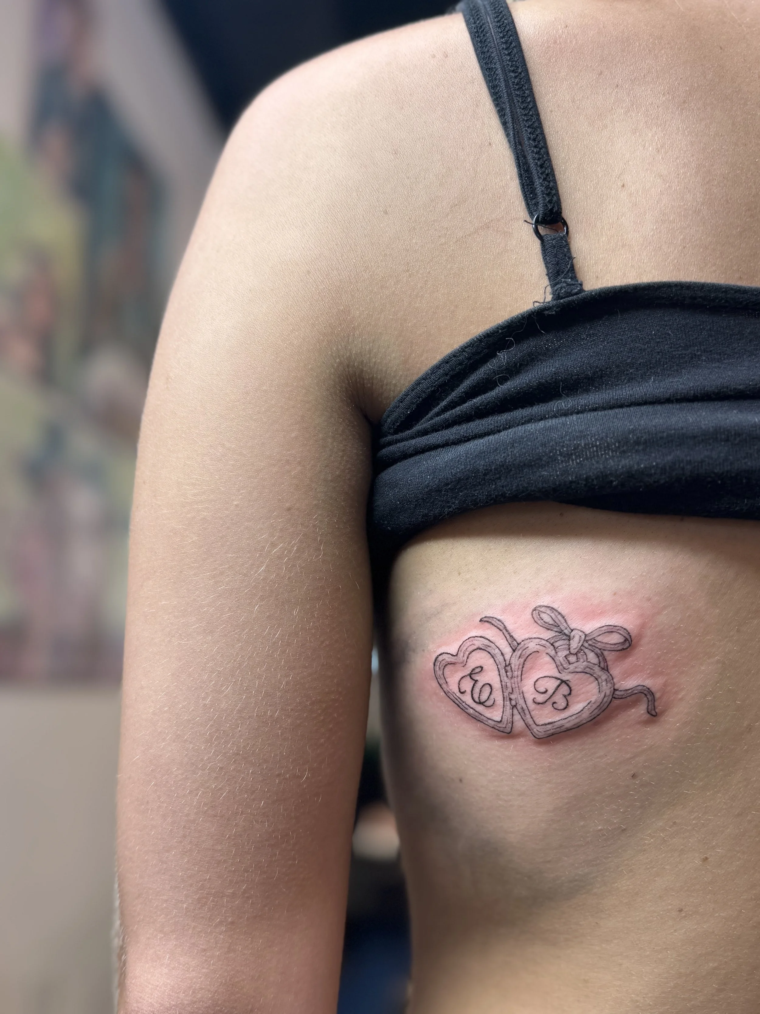 Close-up of a person's shoulder and side torso, showing a tattoo of two interlinked hearts with initials 'E' and 'B' inside each heart, colored in black ink. The person is wearing a dark sleeveless top with an adjustable strap.