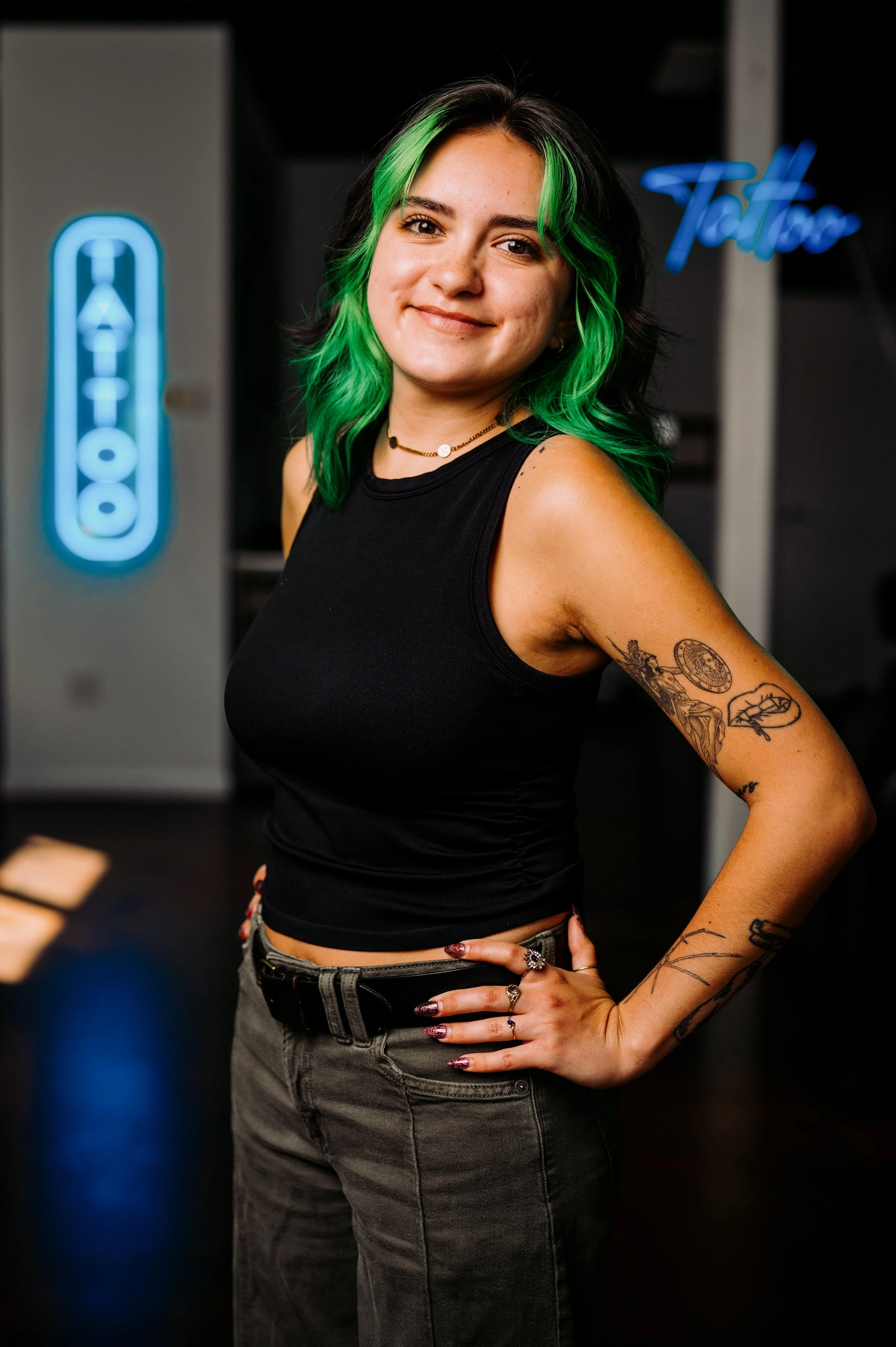 Young woman with green and black hair wearing a black tank top and dark pants, standing with hand on hip, tattoos on arm, smiling, in a room with neon signs and a dark background.