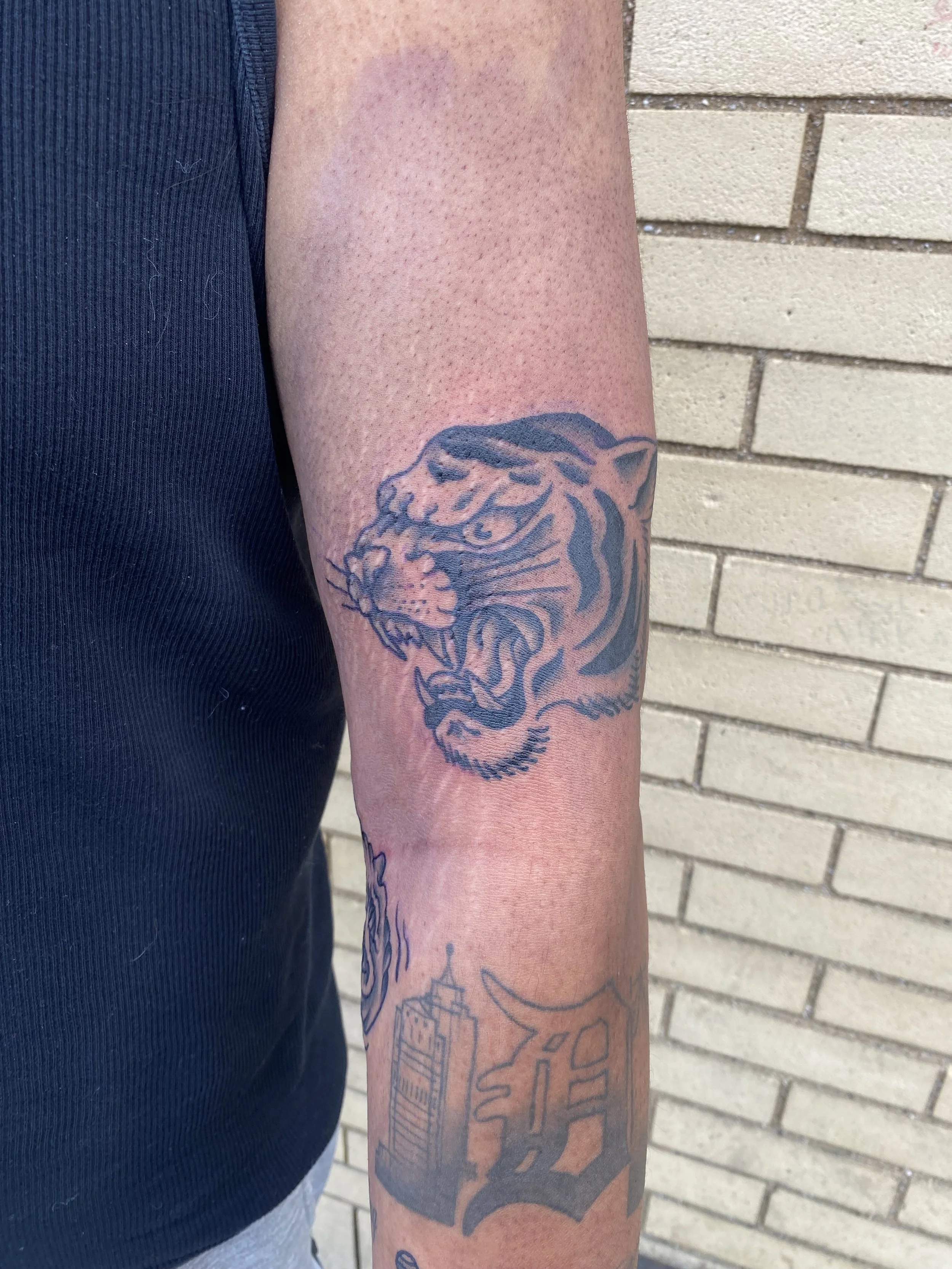 A person with a tattoo of a tiger's head on their forearm, standing against a brick wall. The individual is wearing a dark shirt.