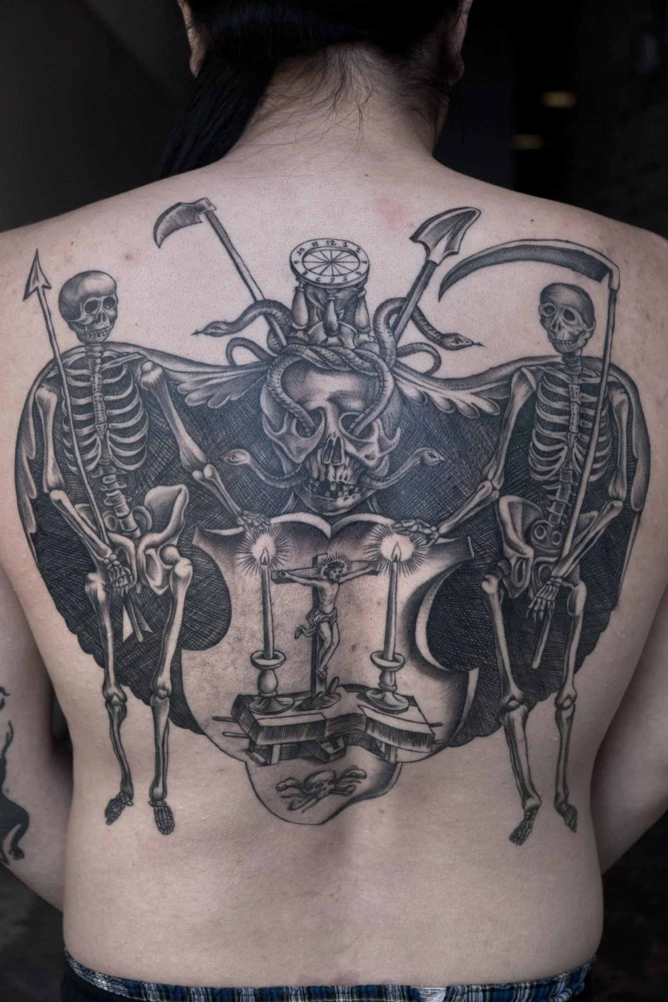 A large tattoo on a person's back featuring various skeletons, skulls, scythes, a crucifix, and other symbolic elements in black ink.