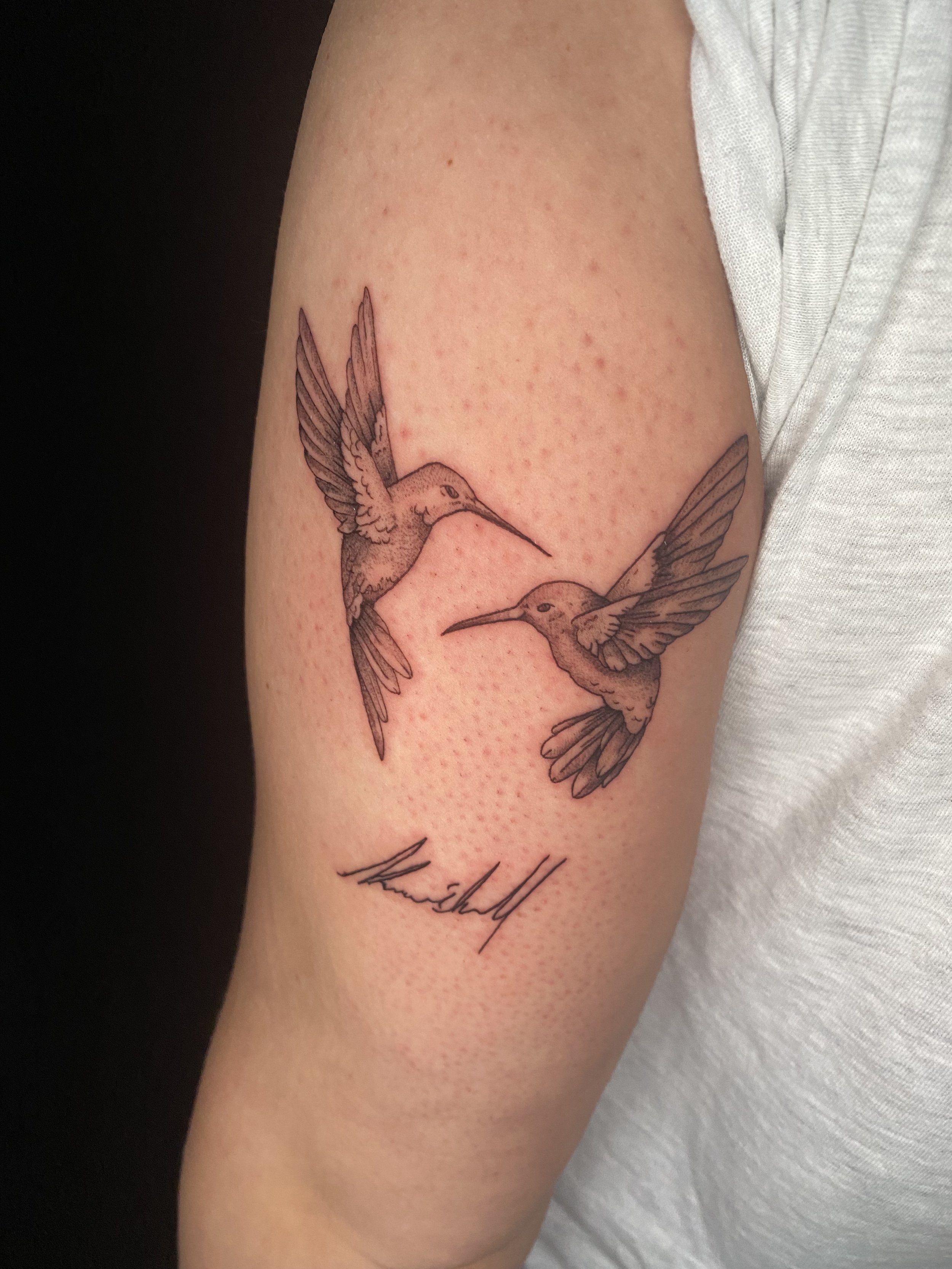 Close-up of a person's upper arm with a tattoo of two hummingbirds, one facing left and the other facing right, with cursive script underneath.