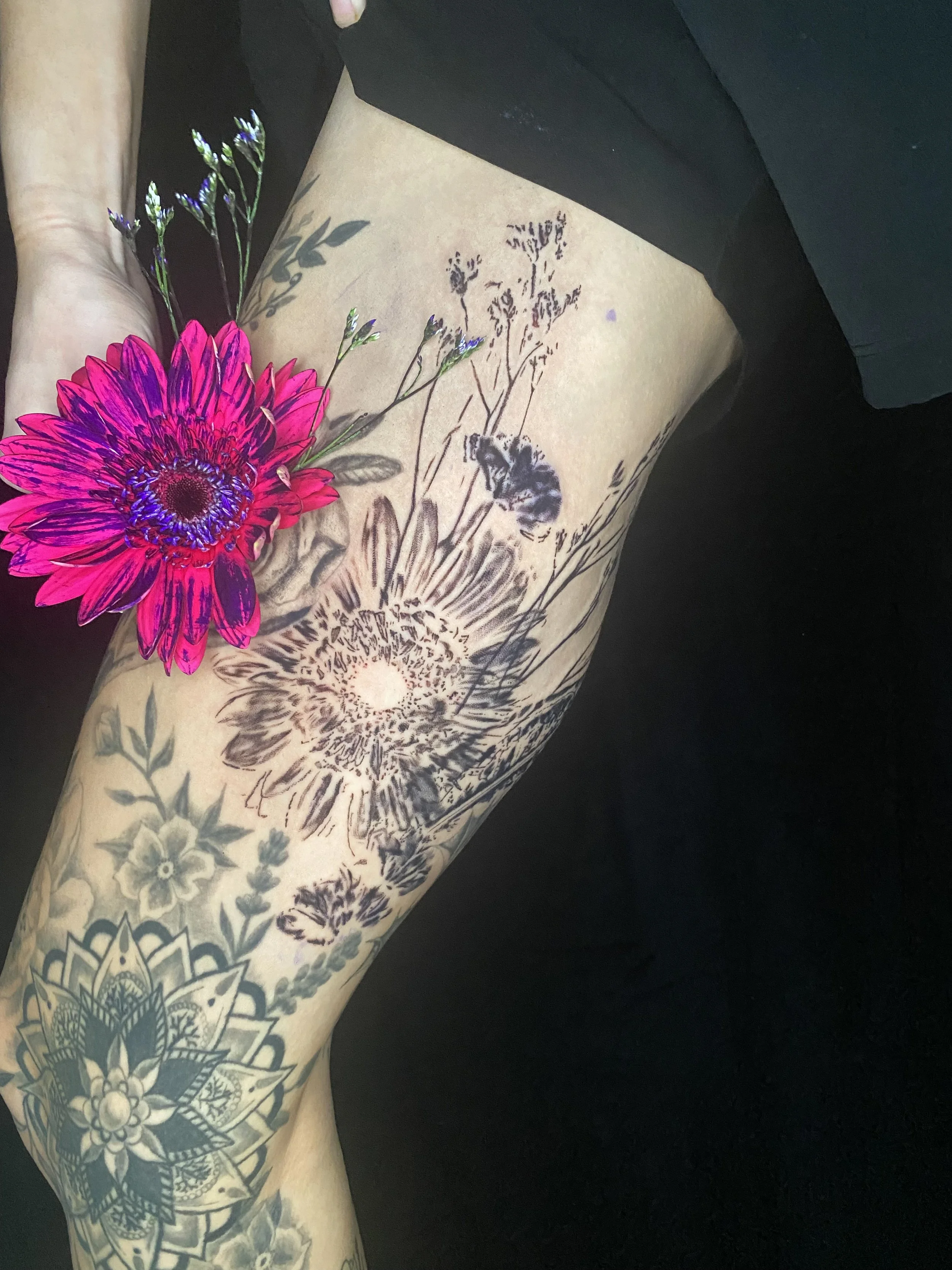 Close-up of a person's arm with detailed black and gray floral tattoos and a large, colorful pink flower placed on the arm.