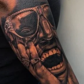 Tattoo of a man's face with sunglasses, holding a gun in his mouth, showing teeth and open mouth