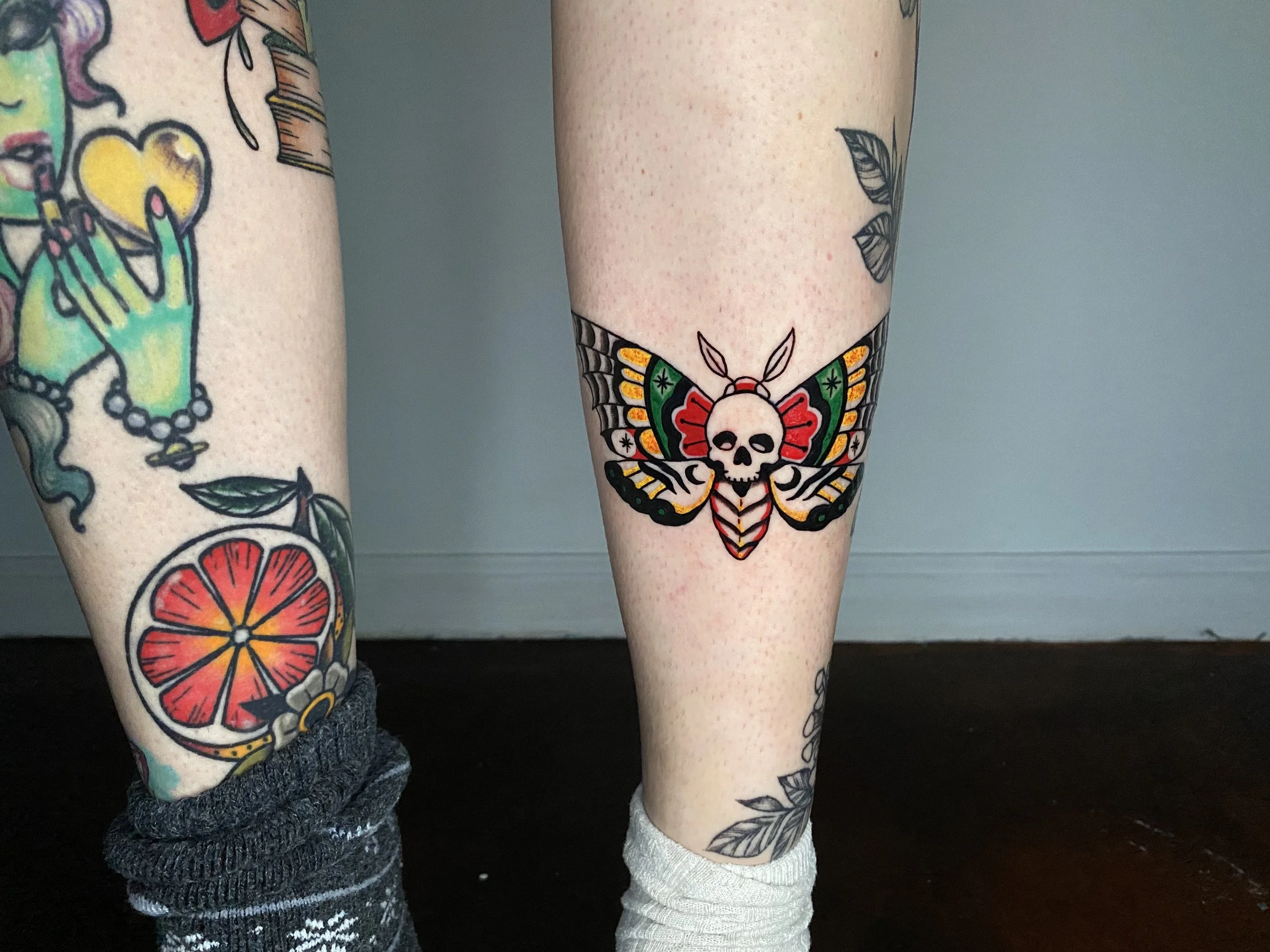 Close-up of two legs with tattoos, one with a colorful skull butterfly and leaf tattoo, and the other with black outlined leaves, both wearing socks and standing on a dark surface.