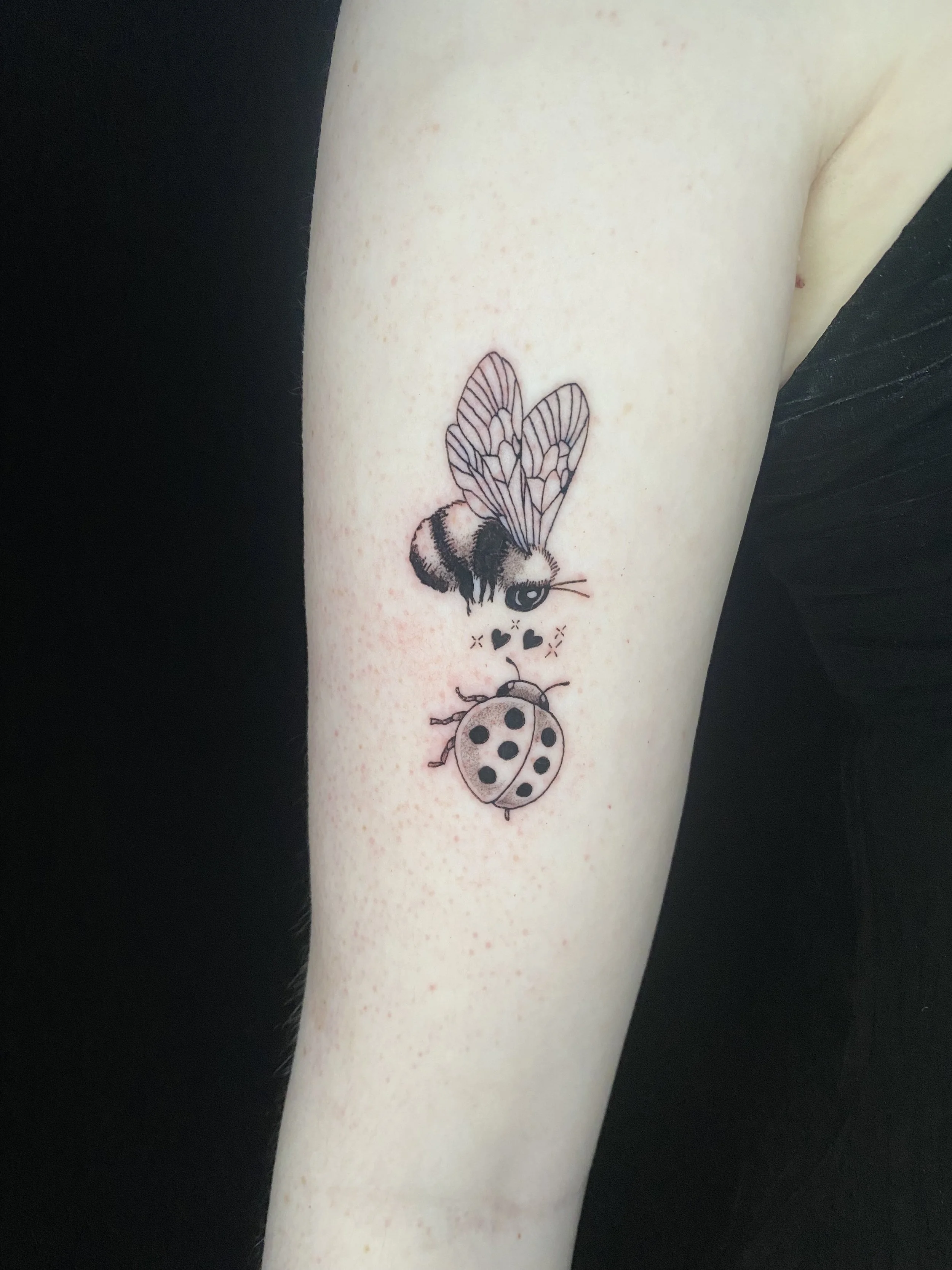 Tattoo of a bee with detailed wings, a ladybug, and small decorative hearts on a person's arm.