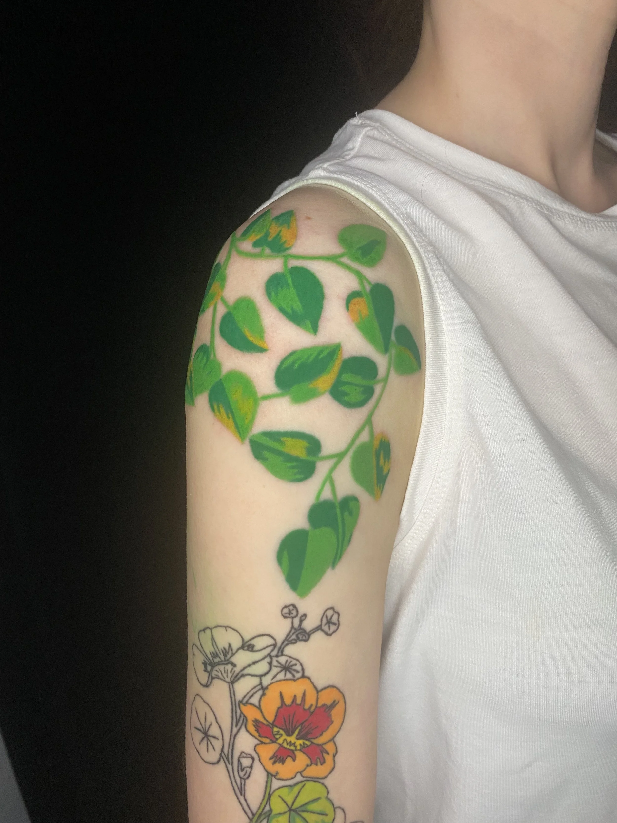 A person's upper arm and shoulder with a colorful flower and leaf tattoo, wearing a white sleeveless shirt, against a black background.