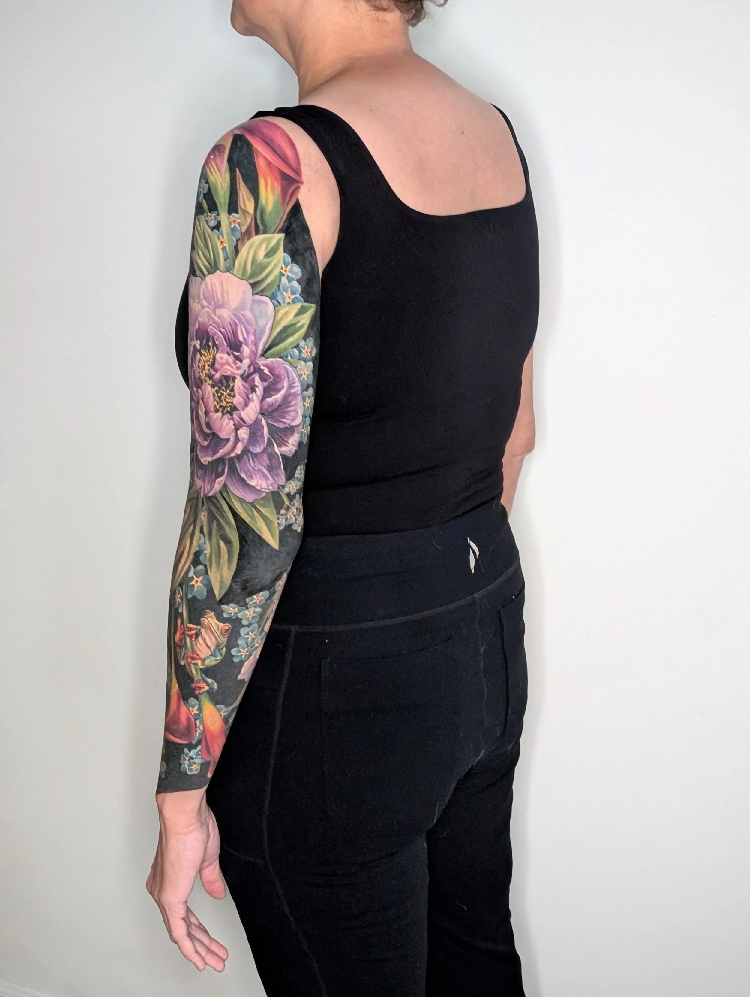 The image shows a person with short, curly hair wearing a black tank top and black pants, standing against a plain white wall. They have a large, colorful floral tattoo covering their entire left arm.