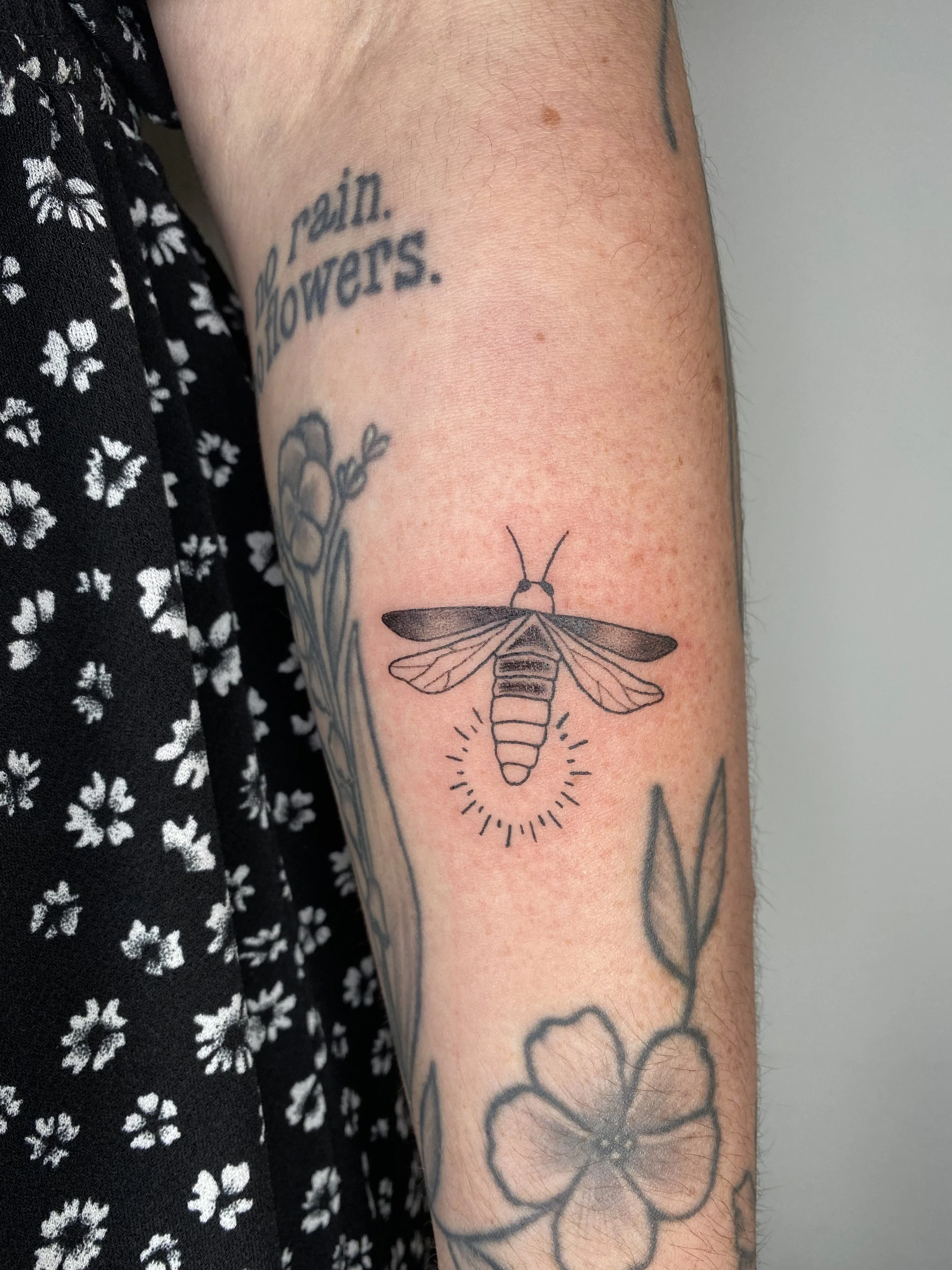 Close-up of a person's arm with a tattoo of a bee and a flower, and a black shirt with white floral pattern.