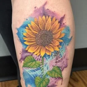 Colorful tattoo of a sunflower with green leaves, blue and purple watercolor background, and white spiderweb details.