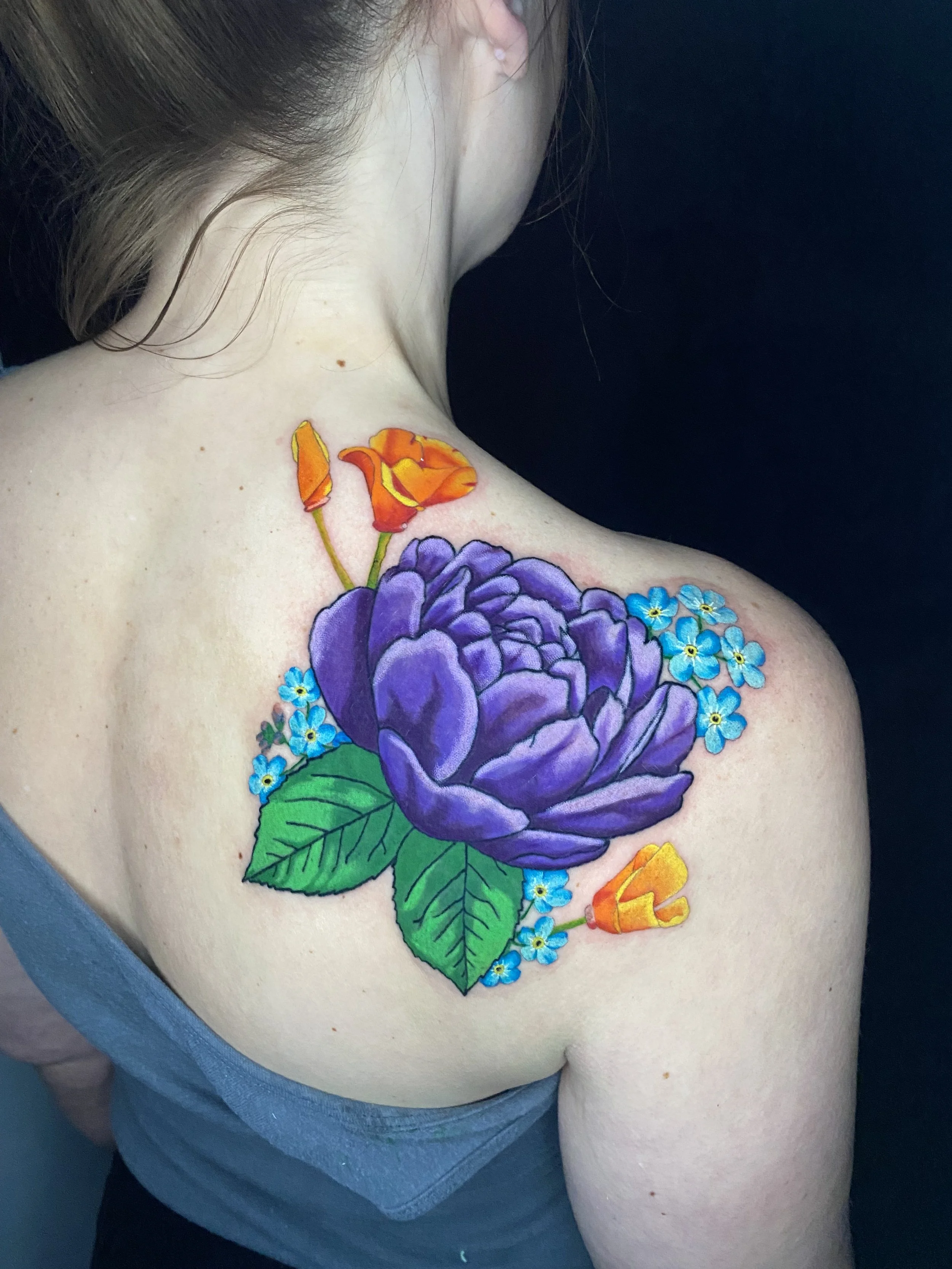 Colorful tattoo of purple flower with orange and yellow buds and small blue flowers on woman's shoulder and upper chest.