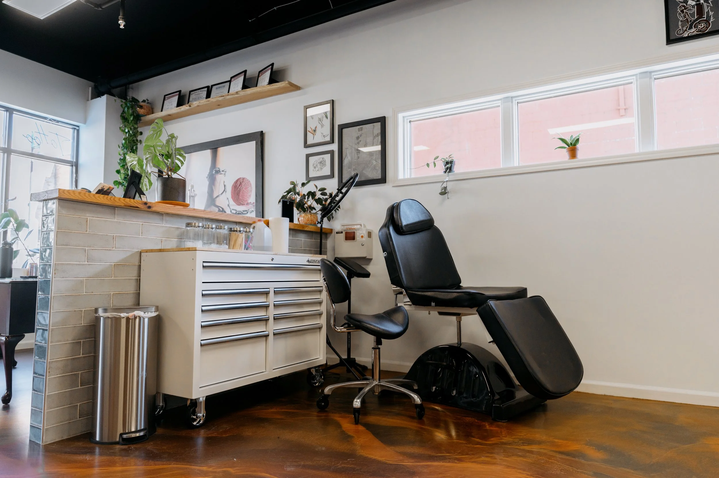 A modern office reception area with a white counter, black ergonomic chairs, potted plants, framed artwork, and windows allowing natural light.