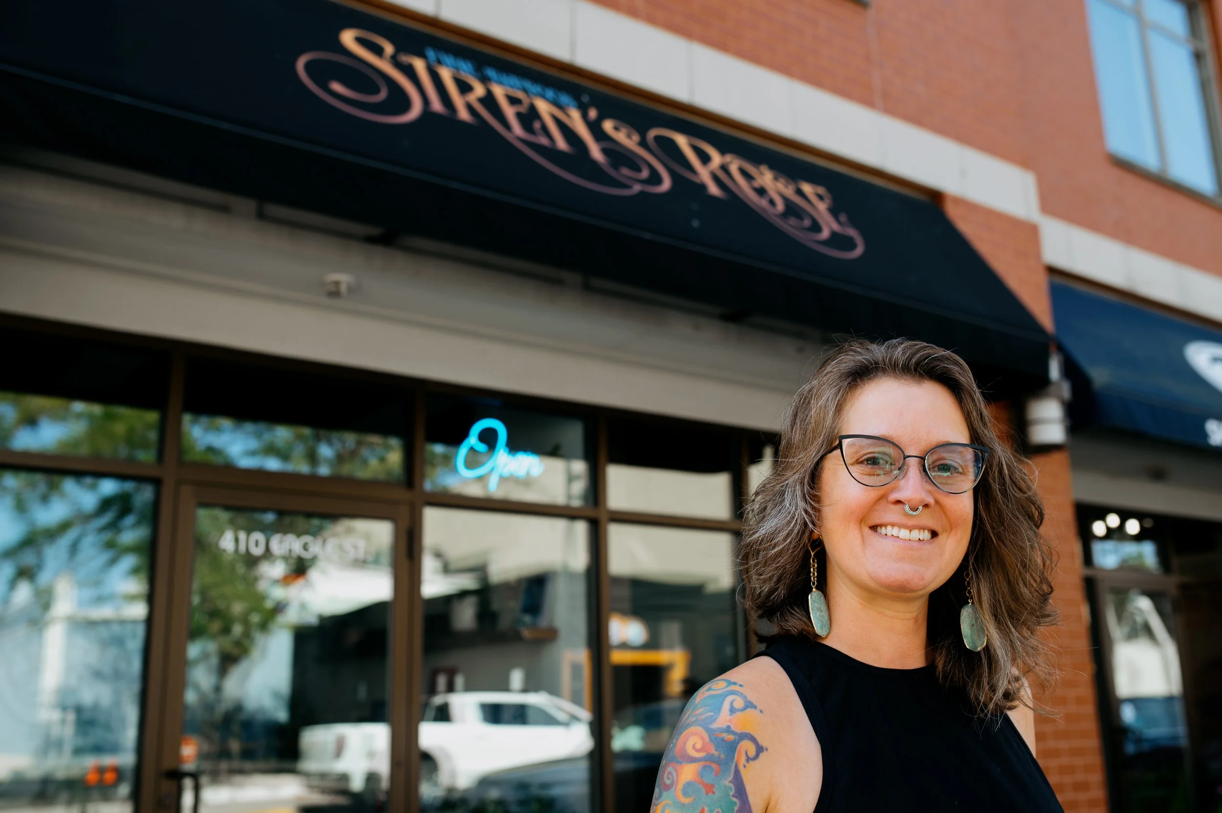 A woman with brown hair, glasses, and large turquoise earrings smiling outdoors in front of a storefront with a black awning that says "Open" and a neon sign also saying "Open."
