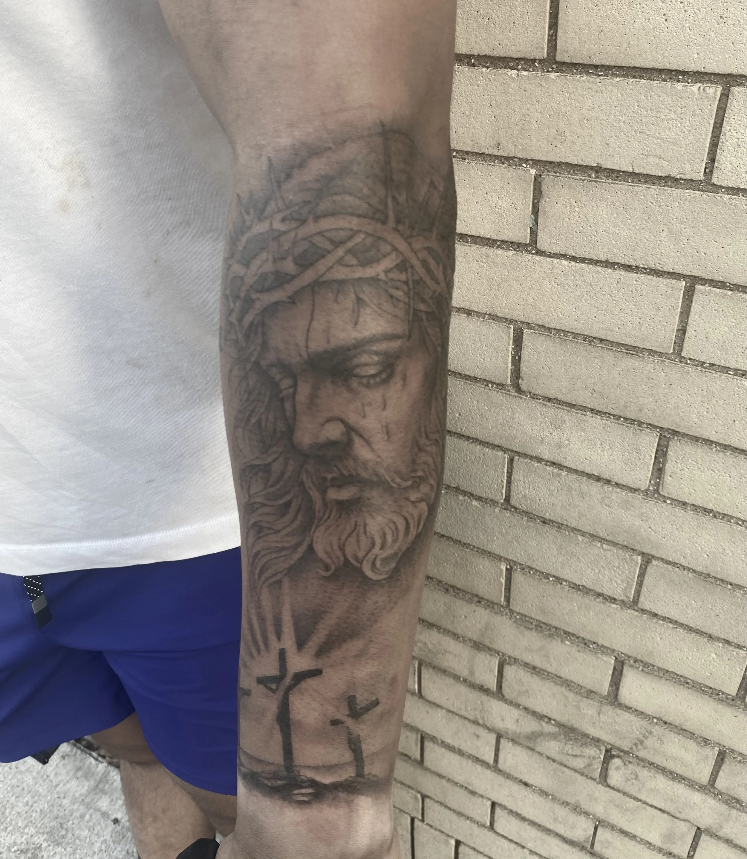 Close-up of a person's arm with a large tattoo featuring a face, trees, and abstract patterns, standing against a brick wall.