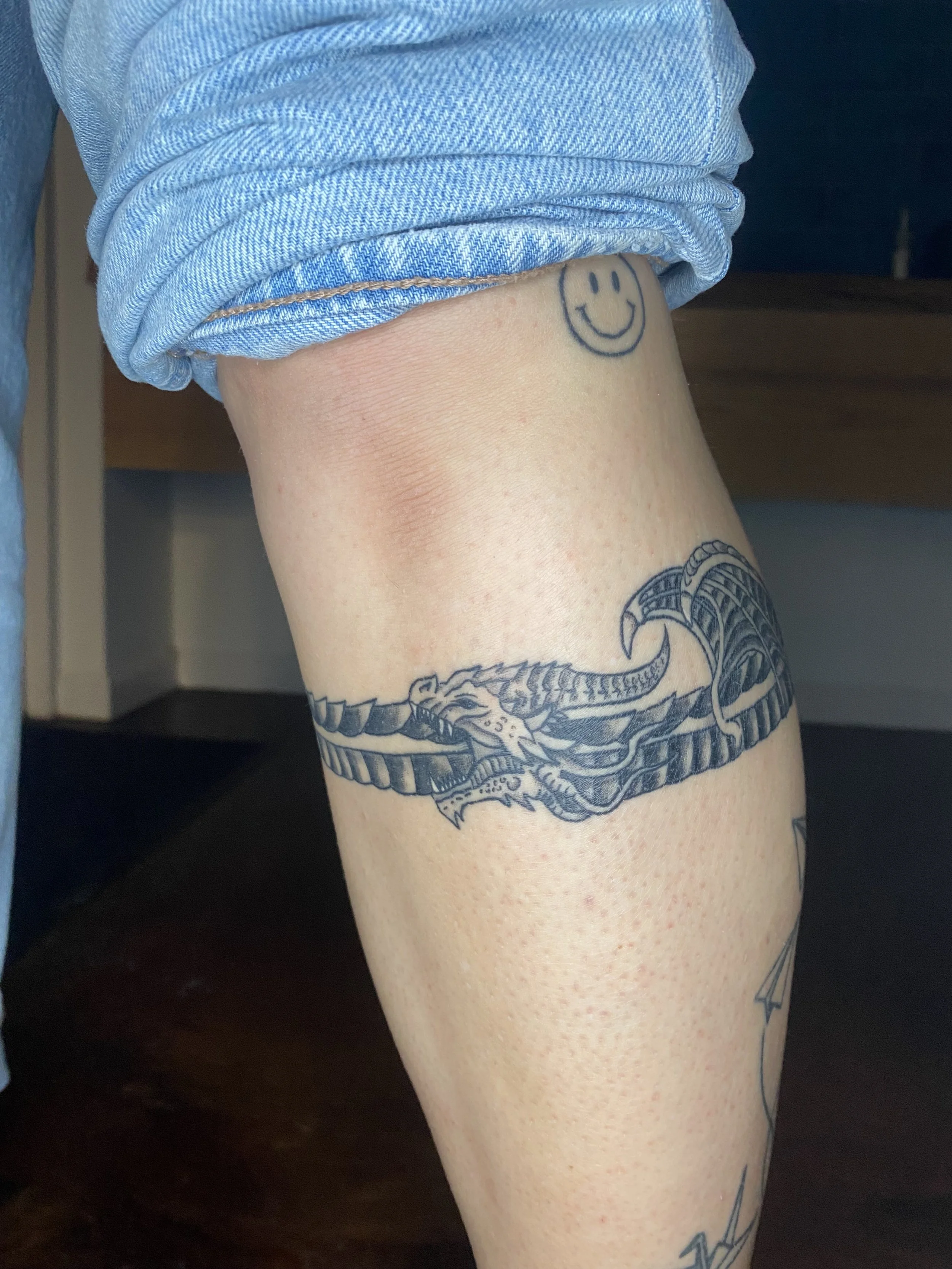 A person's leg with a tattoo of a smiling face on their ankle and a snake with open mouth tattooed around their calf, wearing rolled-up light-wash denim jeans.
